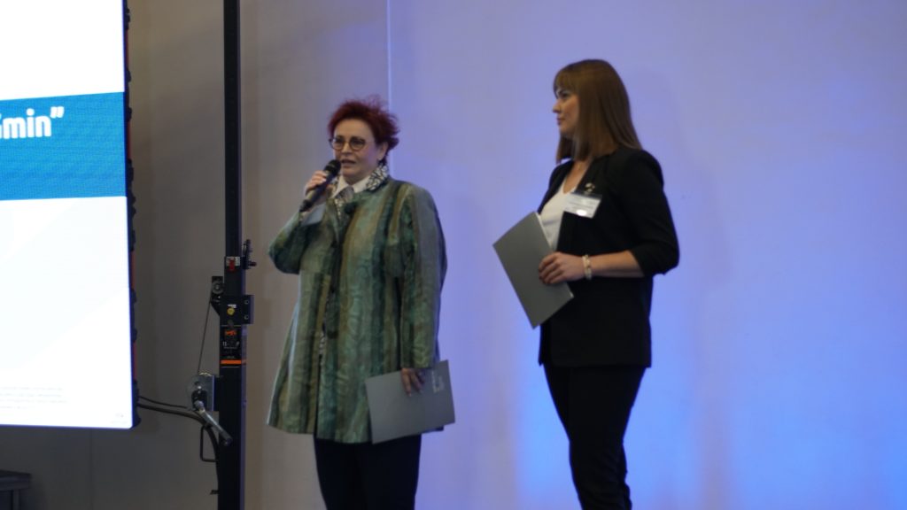 Panie Prezes stoją z teczkami w ręku. Jolanta Ratajczak mówi coś przez mikrofon.