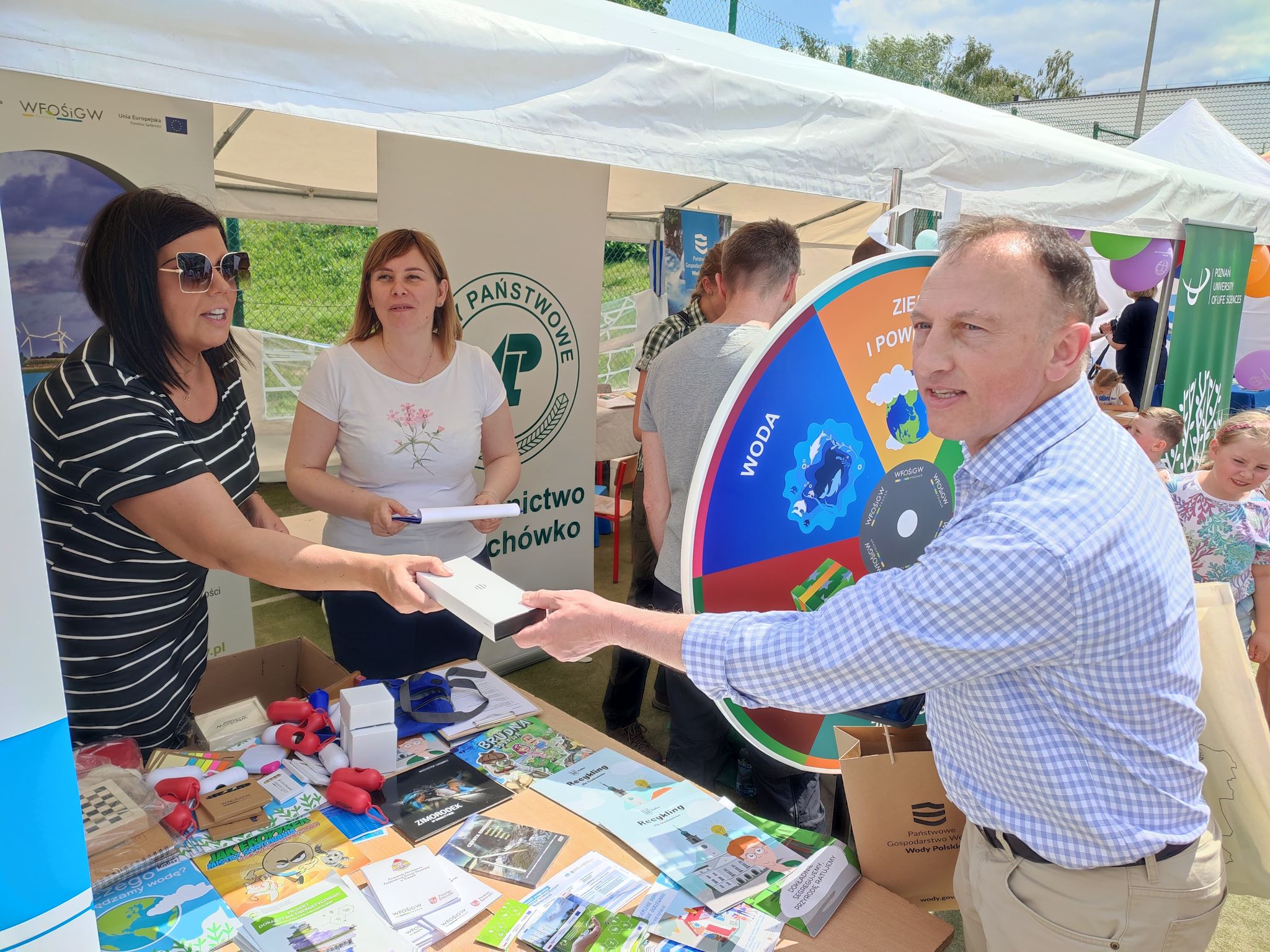 3 osoby wokół stoiska wystawienniczego podczas festynu.