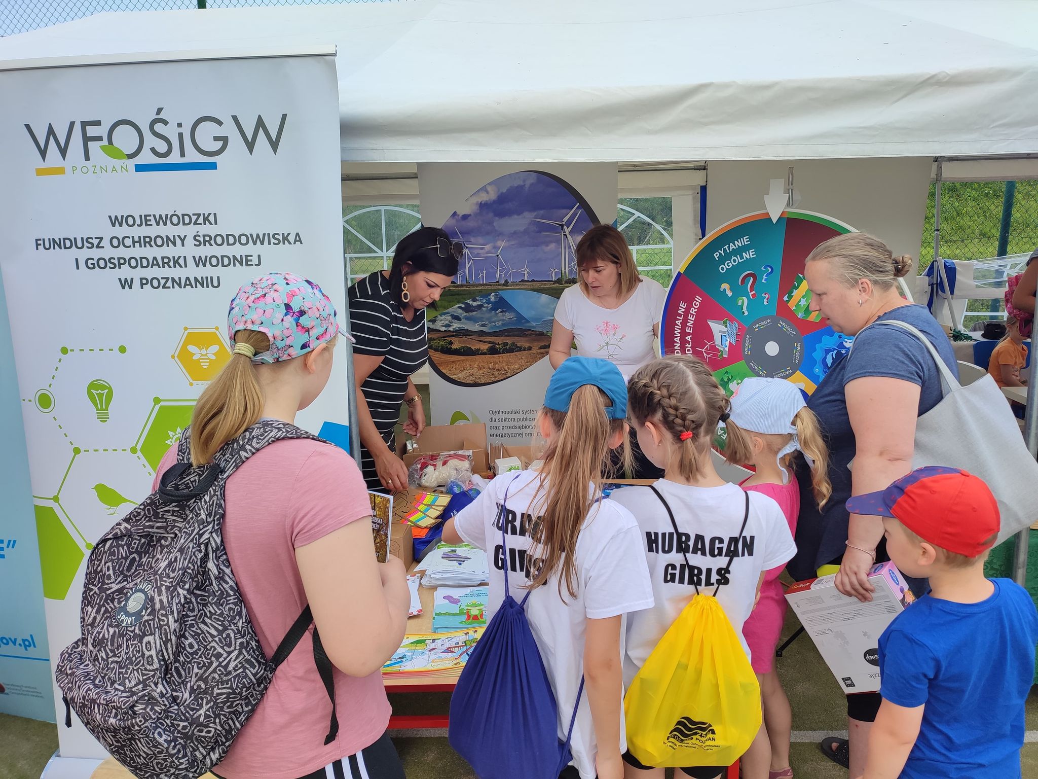 Grupa osób wokół stoiska wystawienniczego podczas festynu.