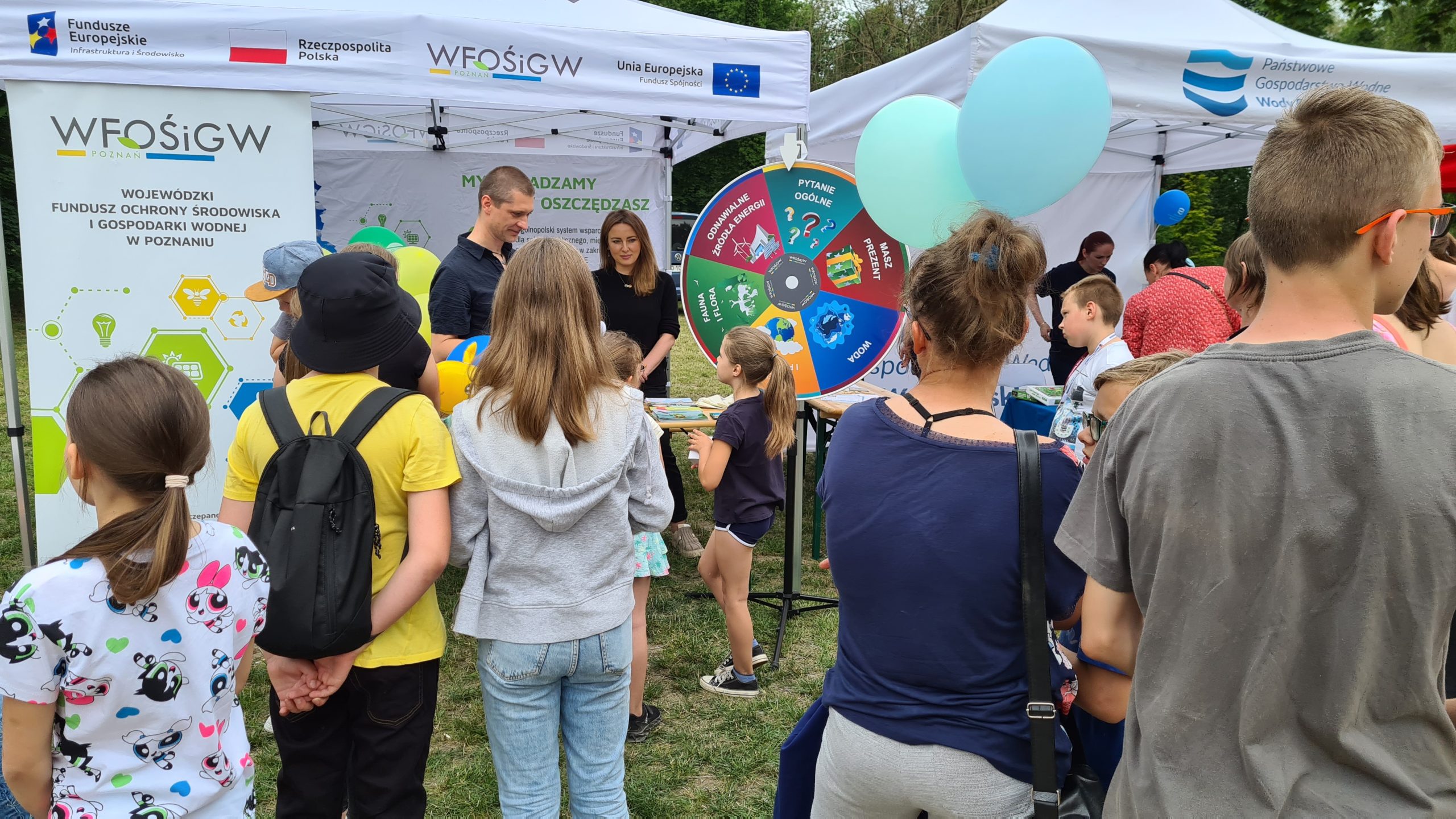 Grupa ludzi na tle stoiska w trakcie pikniku.