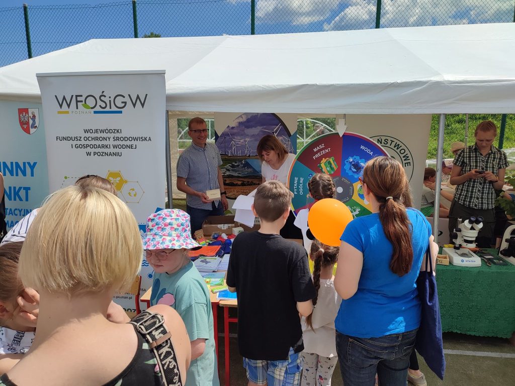 Grupa osób wokół stoiska wystawienniczego podczas festynu.