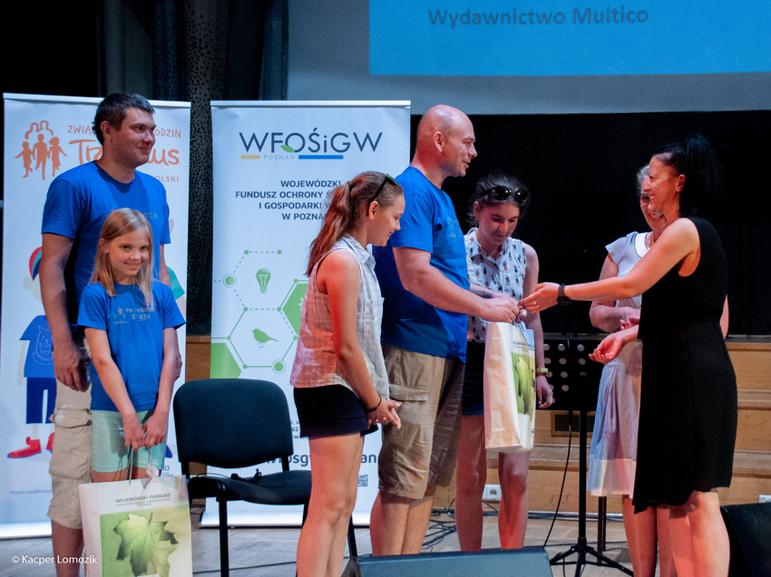 7 osób stoi na scenie na tle bannerów reklamowych.