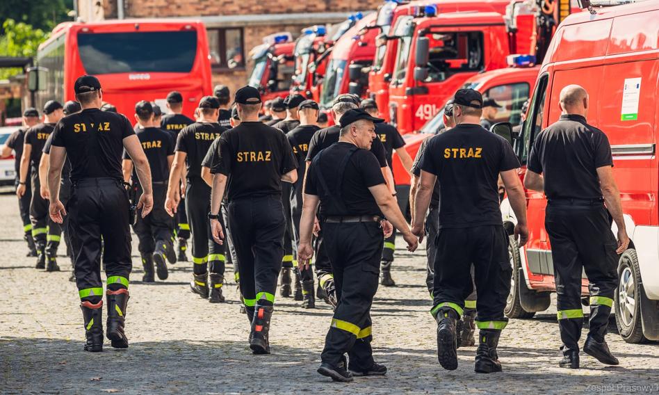 Duża grupa strażaków przy samochodach strażackich.