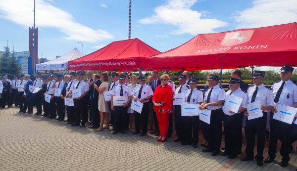 Grupa strażaków w mundurach stoi na placu przed komendą staży pożarnej.