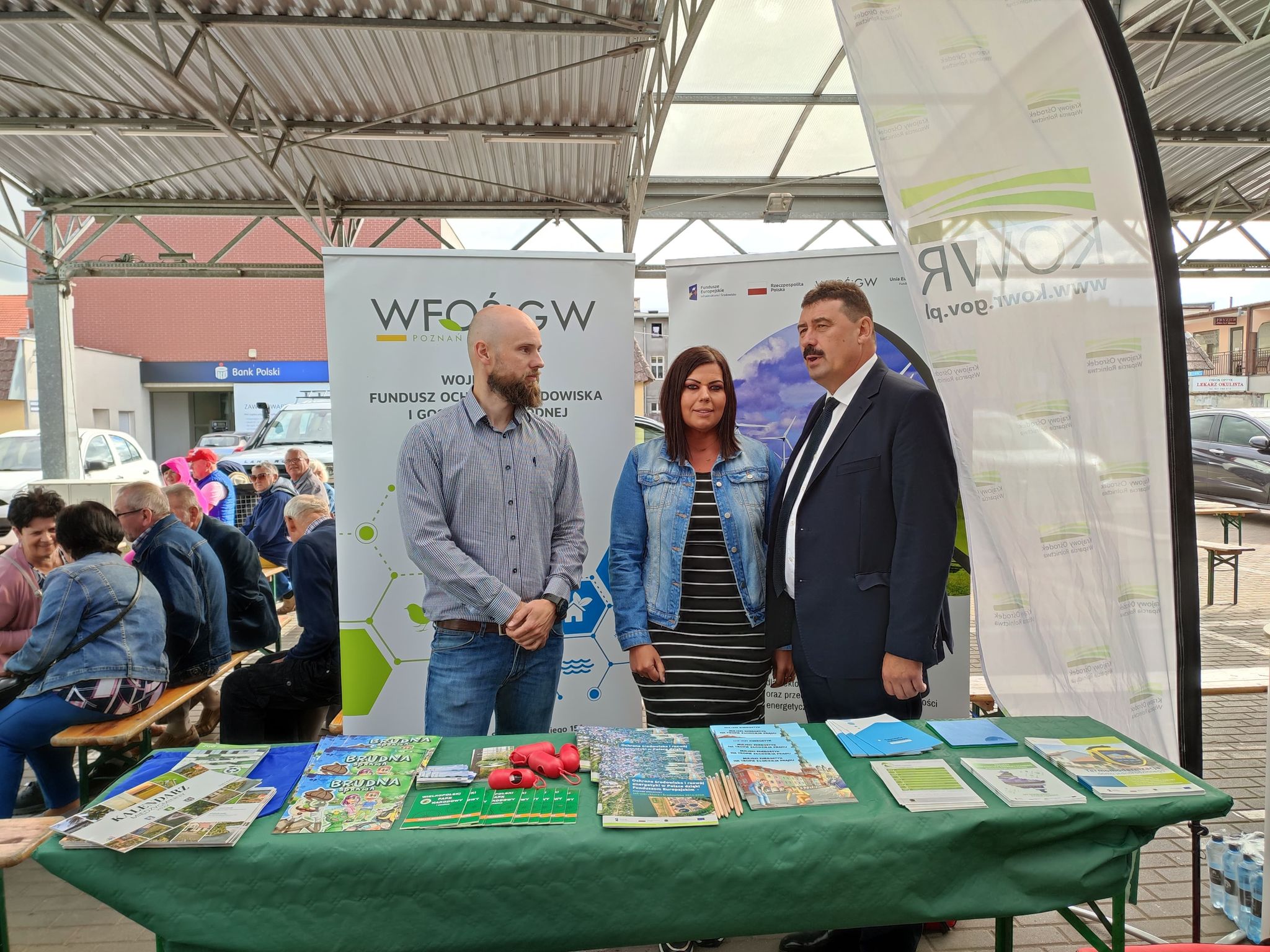 3 osoby stoją przy stoisku w trakcie festynu.