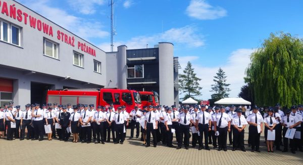 Grupa strażaków w mundurach stoi na placu przed komendą staży pożarnej.