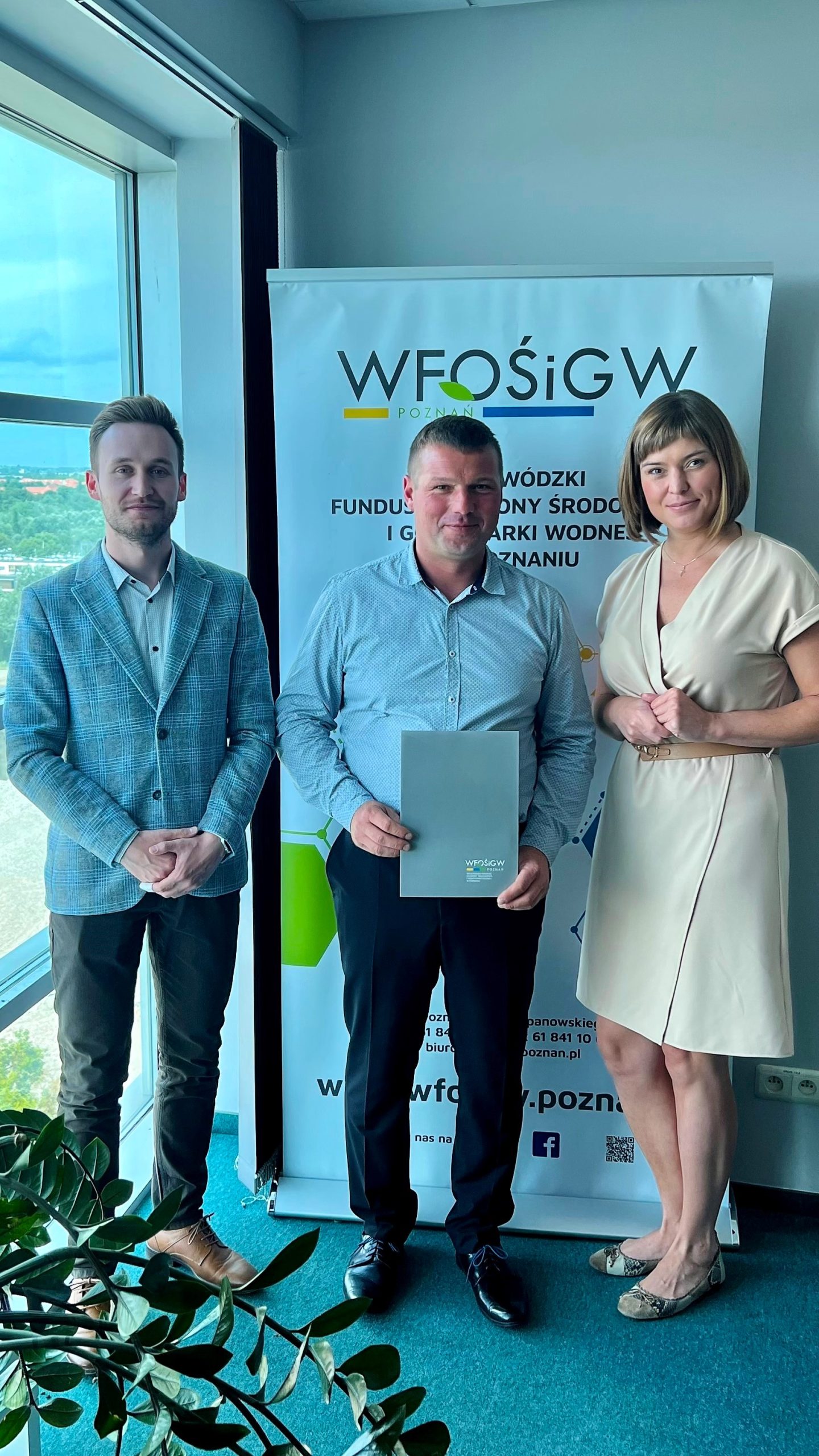 3 osoby stoją na tle ściany i okna.