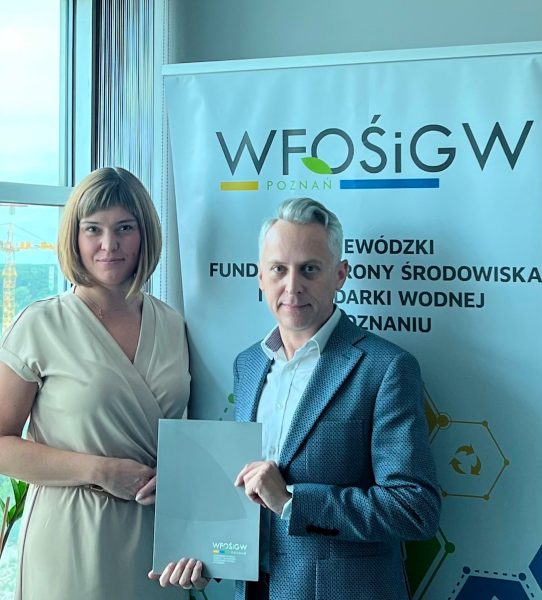 2 osoby stoją na tlę ściany i okna.