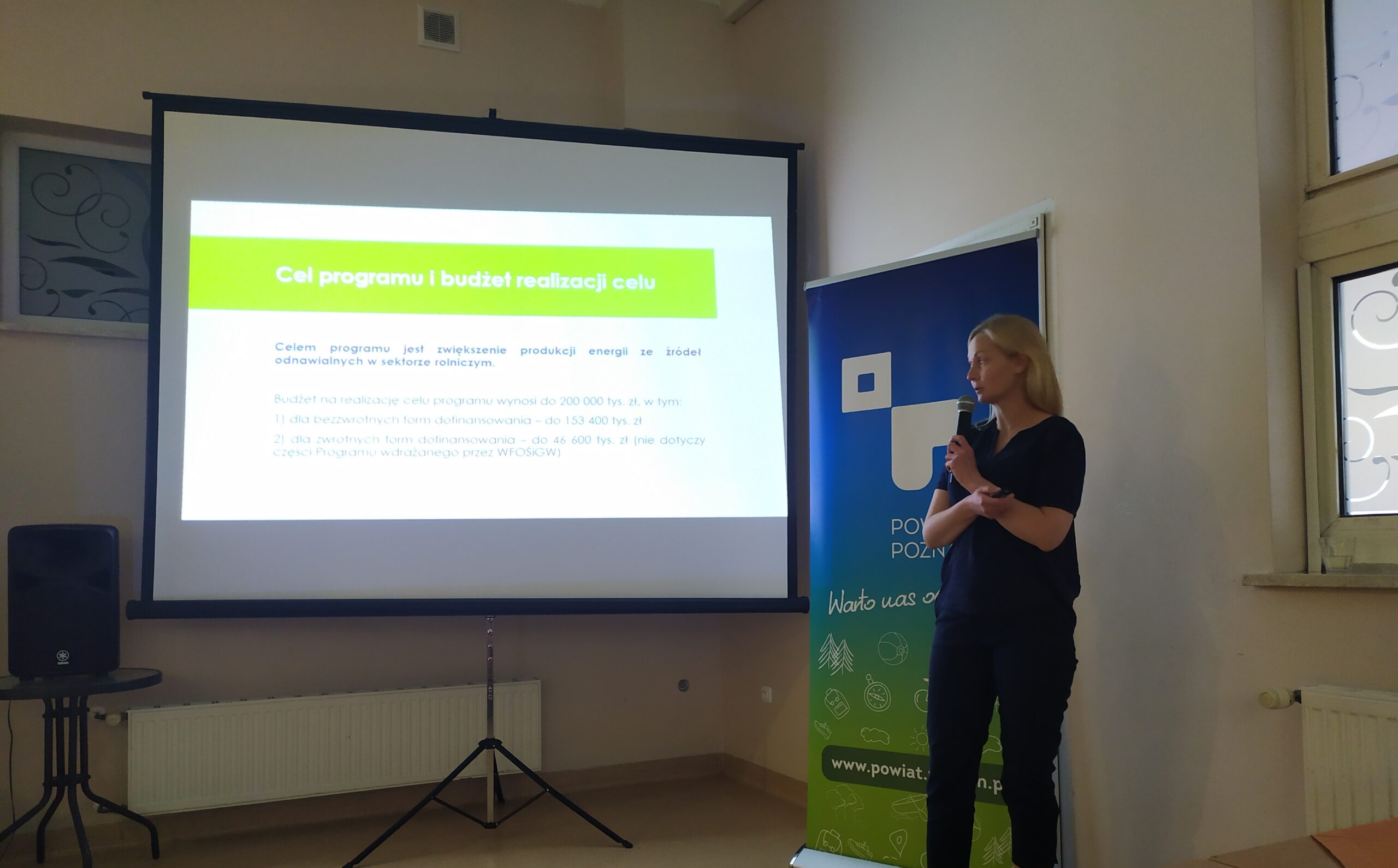 1 osoba stoi na tle przedstawiającym prezentację multimedialną.