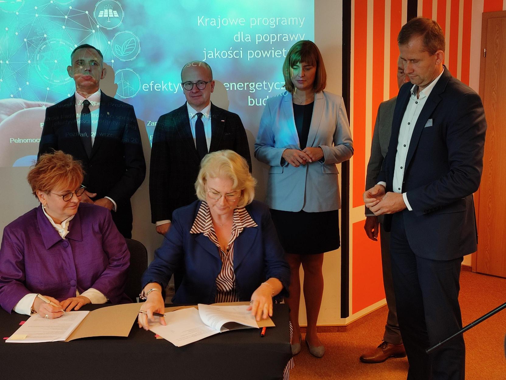 2 osoby siedzą za stołem, a 4 osoby stoją za nimi na tle ściany.