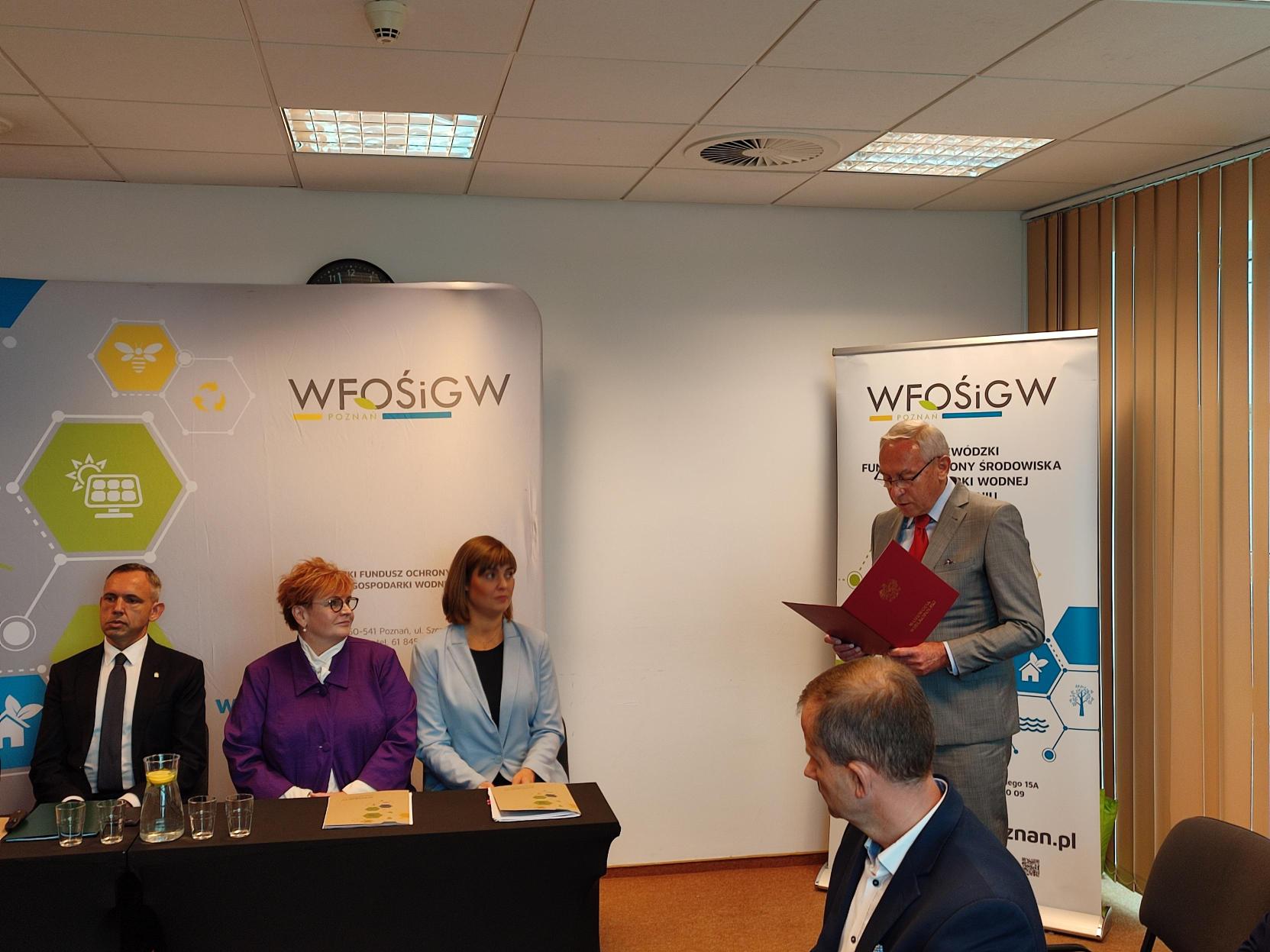 3 osoby siedzą za stołem, a jedna stoi przy ścianie i przemawia.