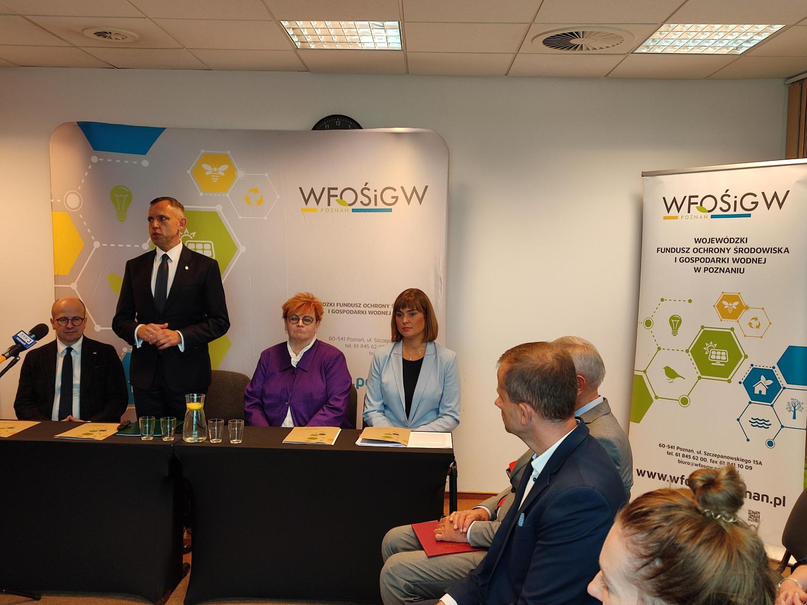 3 osoby siedzą za stołem, a jedna stoi i przemawia.