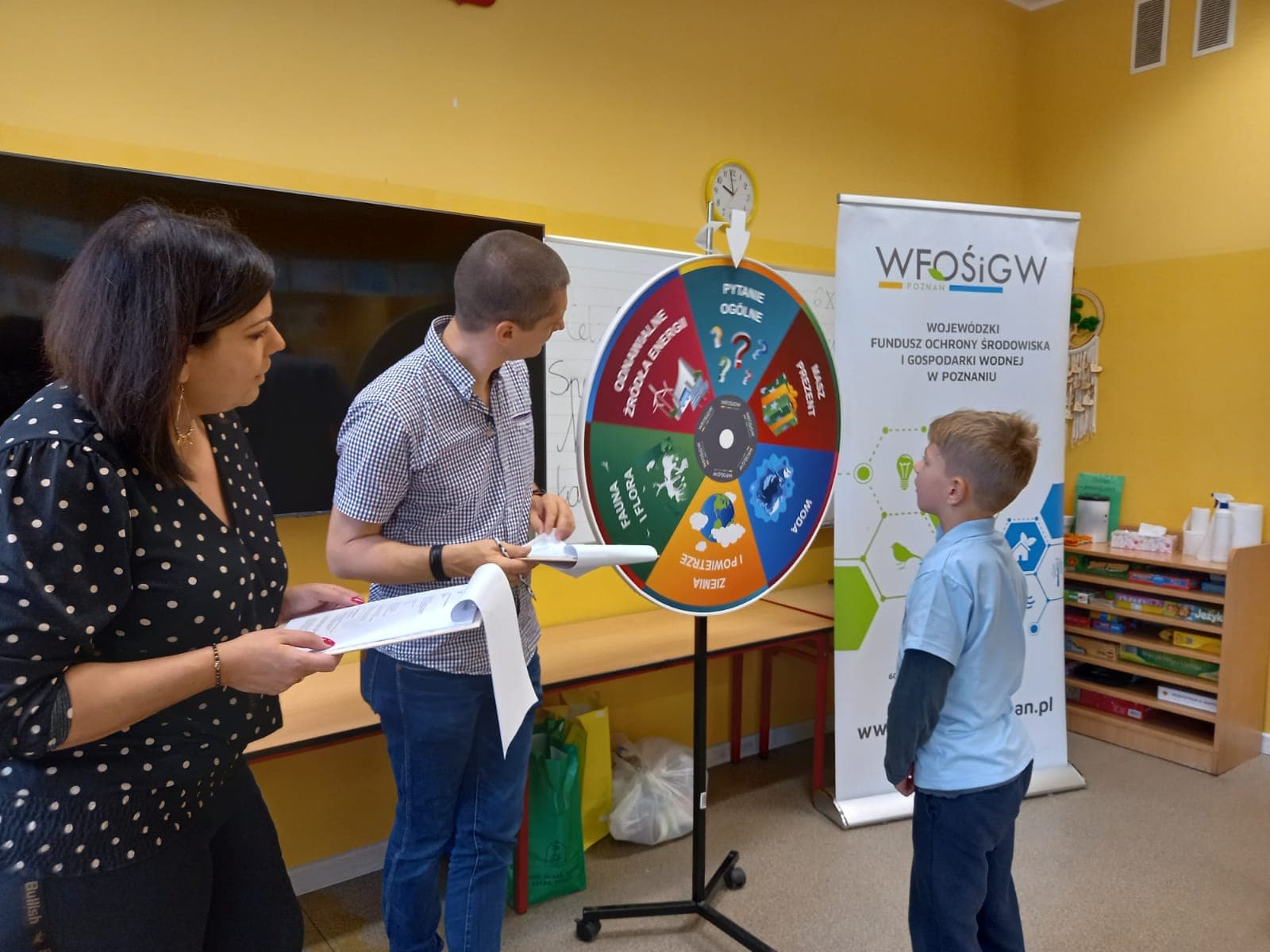 3 osoby stoją na tle ściany i tablicy szkolnej.