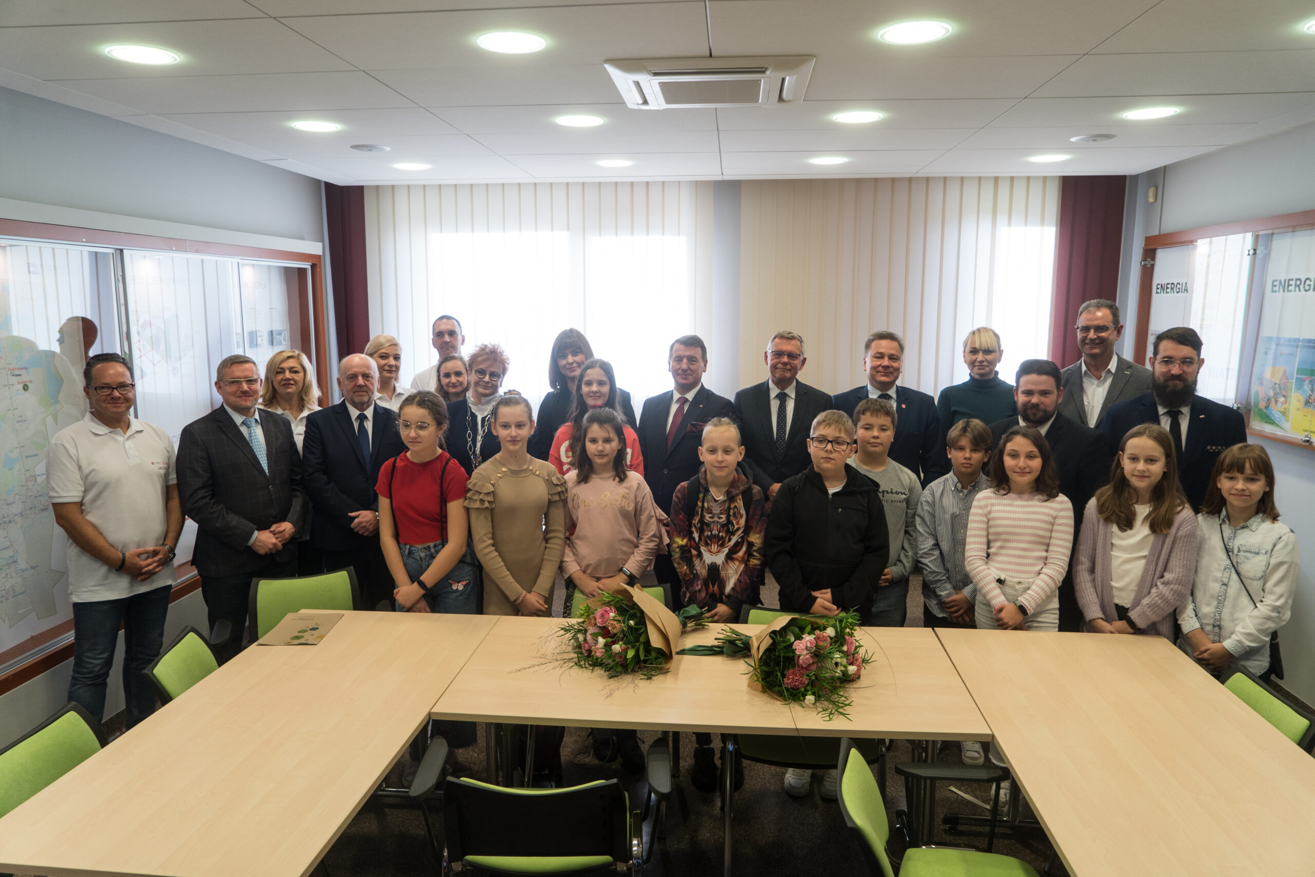 Duża grupa osób stoi na tle okien.