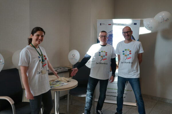 3 osoby stoją na tle ściany obok stołu.