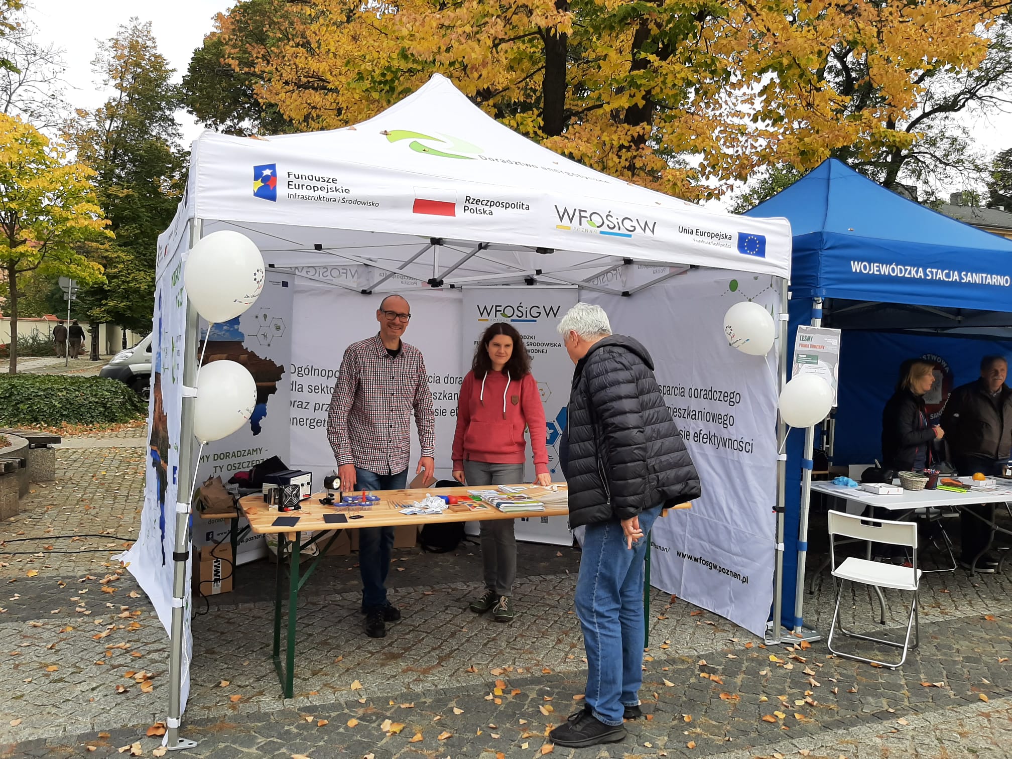 3 osoby stoją przy stoiku podczas festynu.