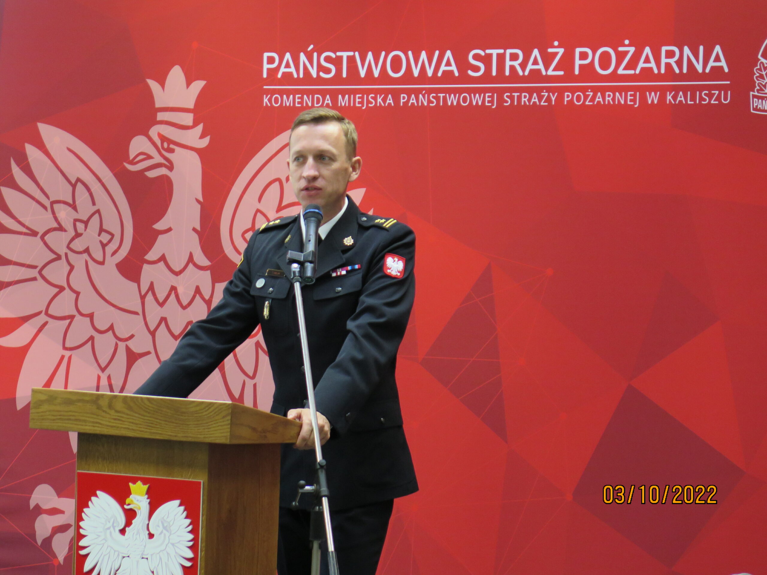 1 osoba stoi na tle ściany przy mównicy.