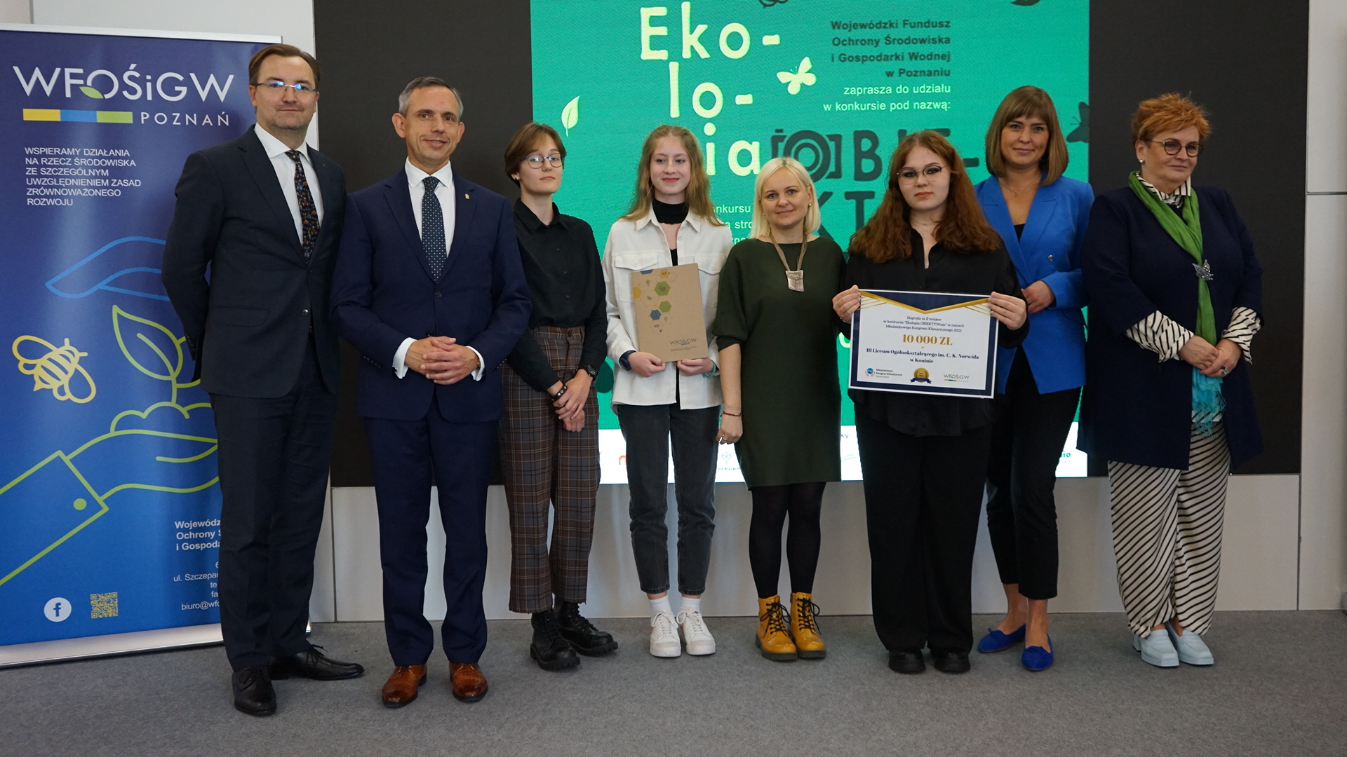 8 osób stoi na tle ściany.