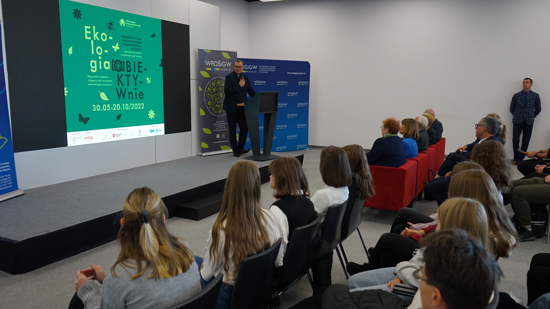 Jedna osoba stoi na scenie przy mównicy.