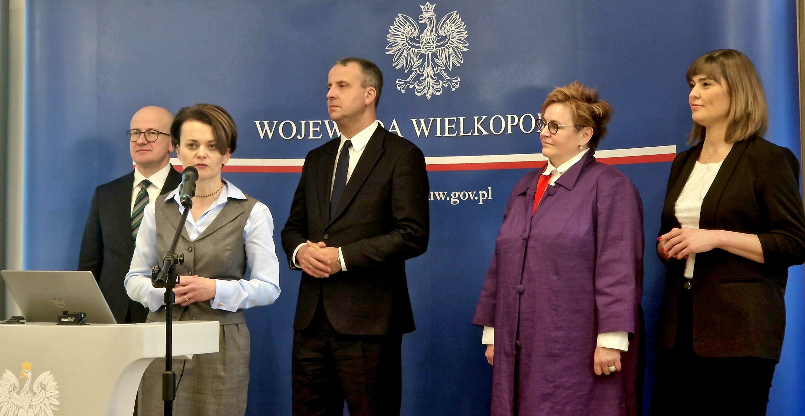 Na zdjęciu jedna osoba stoi przy mównicy, a pozostałe 4 osoby stoją w tle.