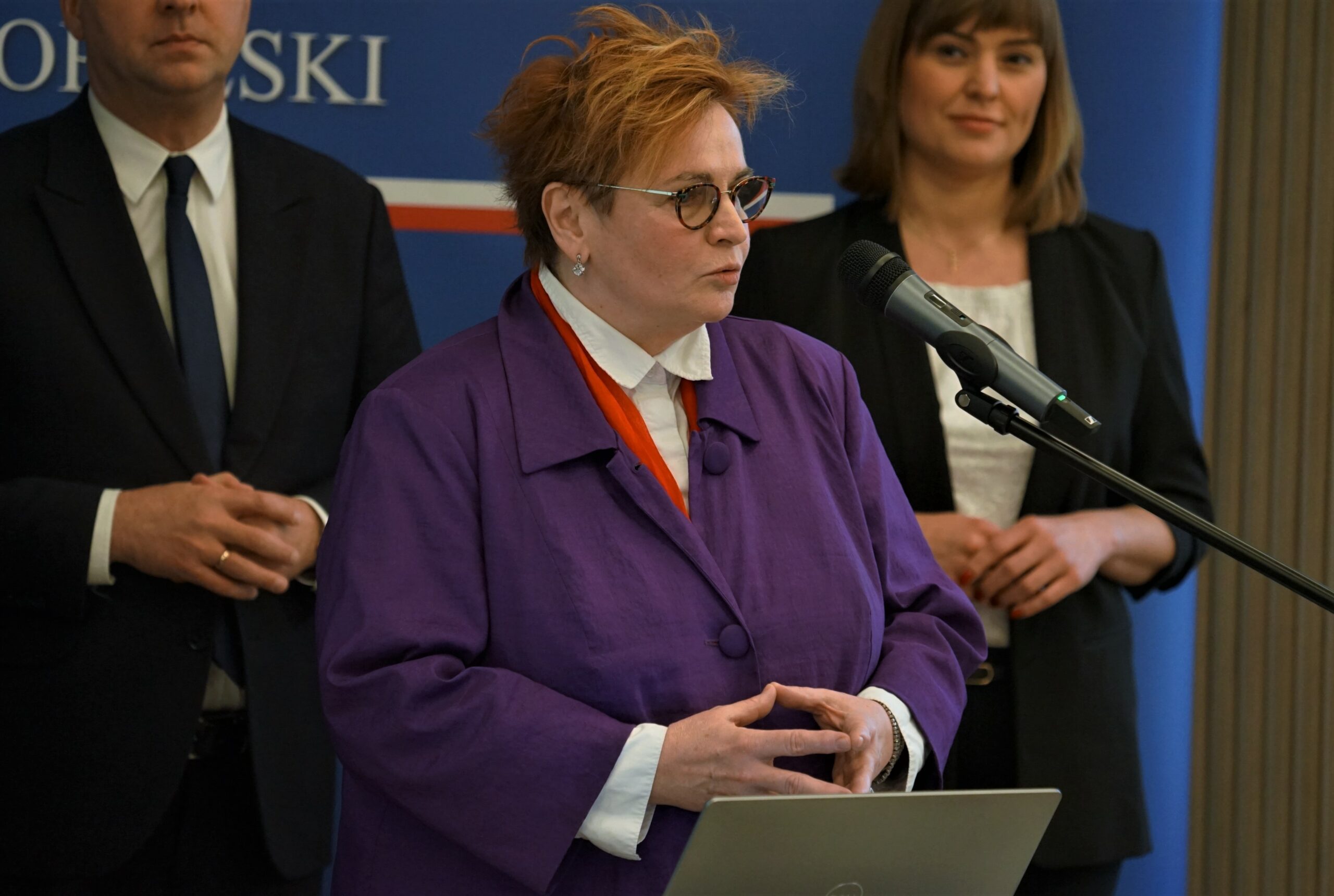 Na zdjęciu osoba na pierwszym planie stoi przy mównicy. Za nią znajdują się w tle jeszcze 2 osoby.