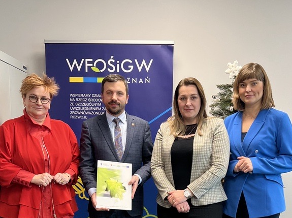 Na zdjęciu stoją 4 osoby na tle ściany i bannera reklamowego.