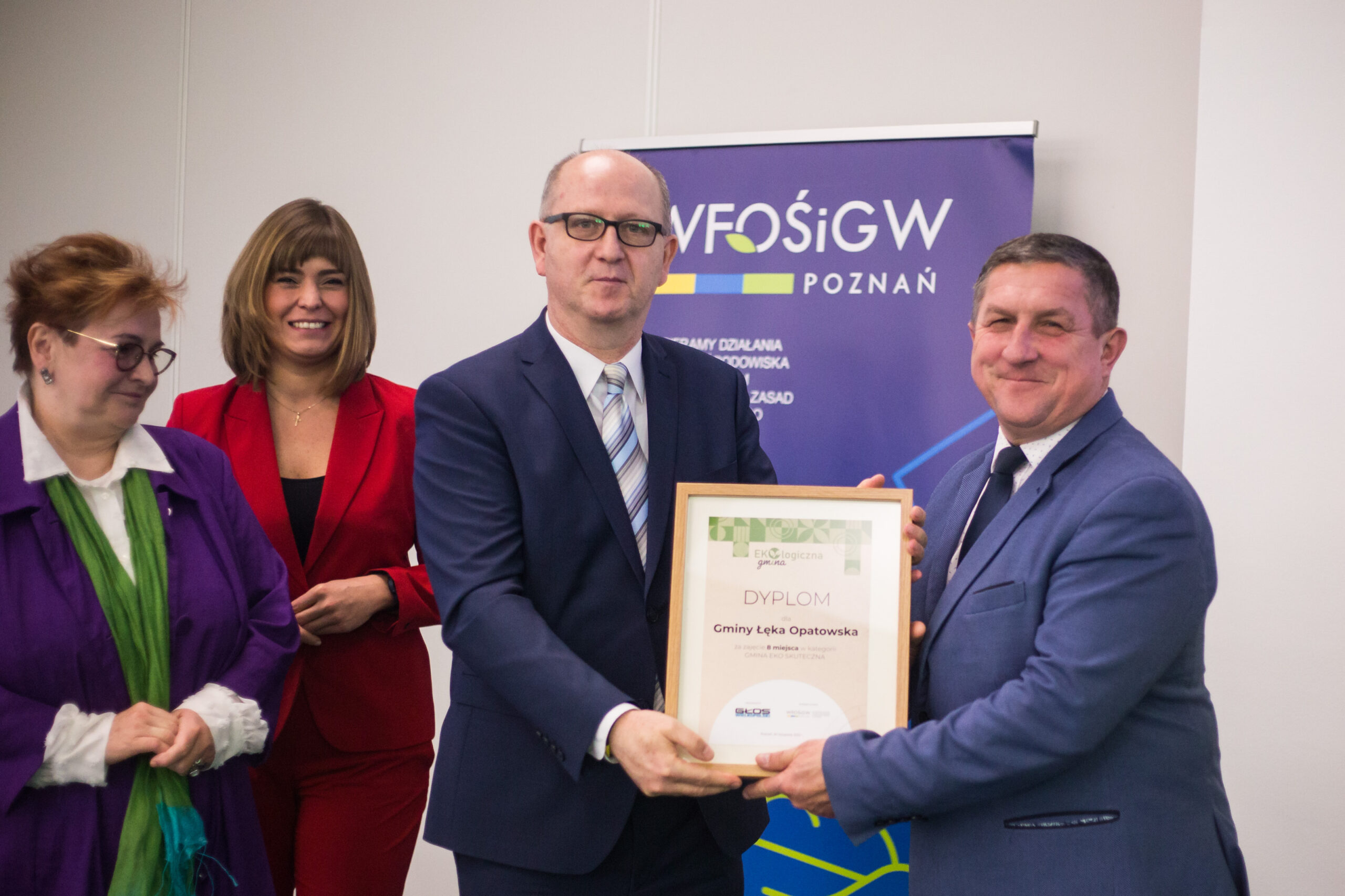 4 osoby stoja na tle bannera reklamowego.