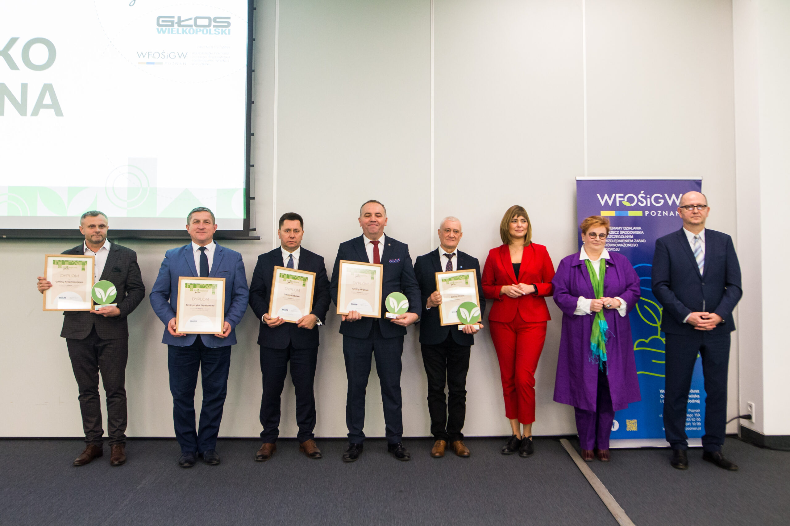 8 osób stoi na tle ściany.