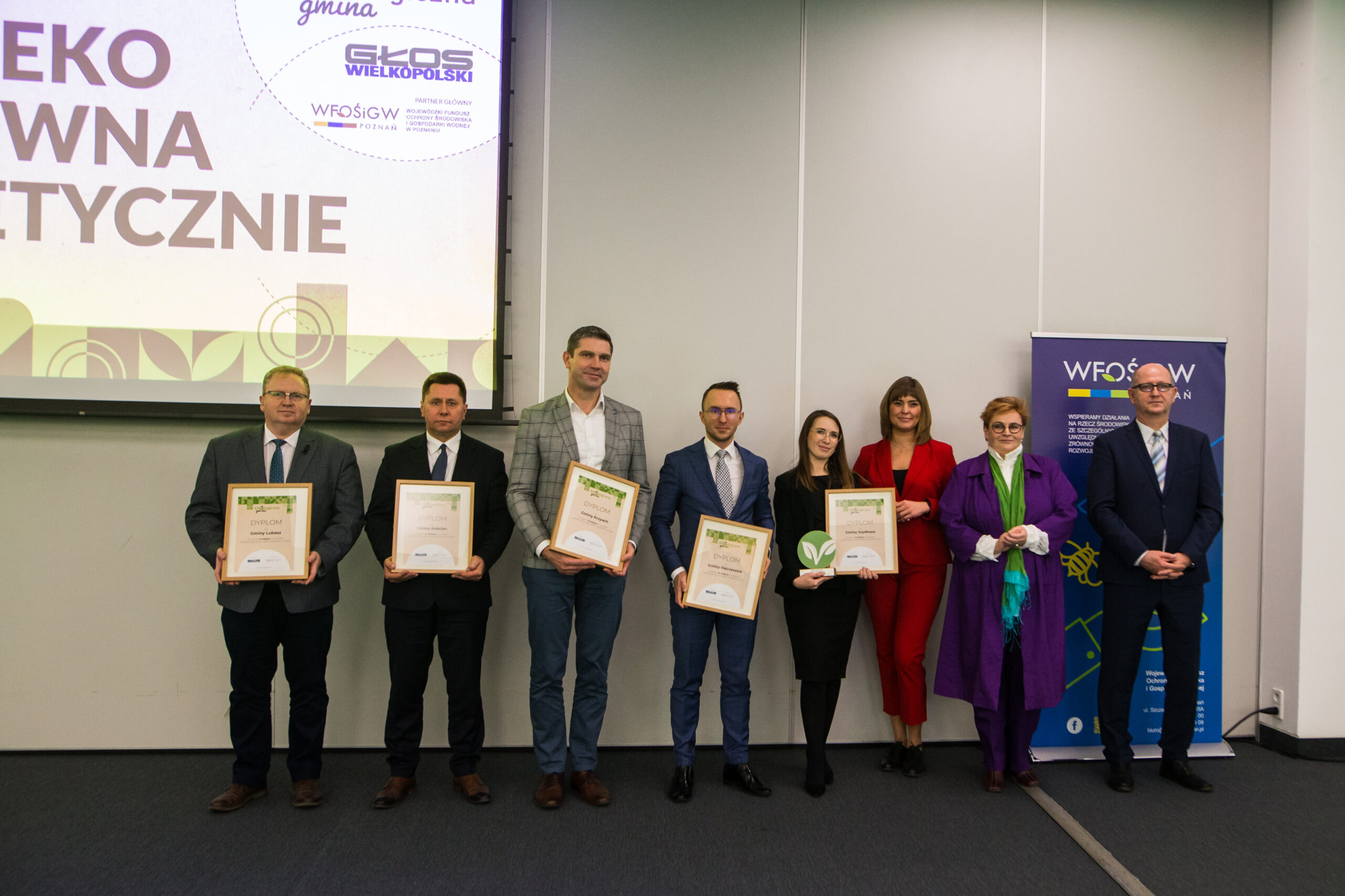 8 osób stoi na tle ściany.