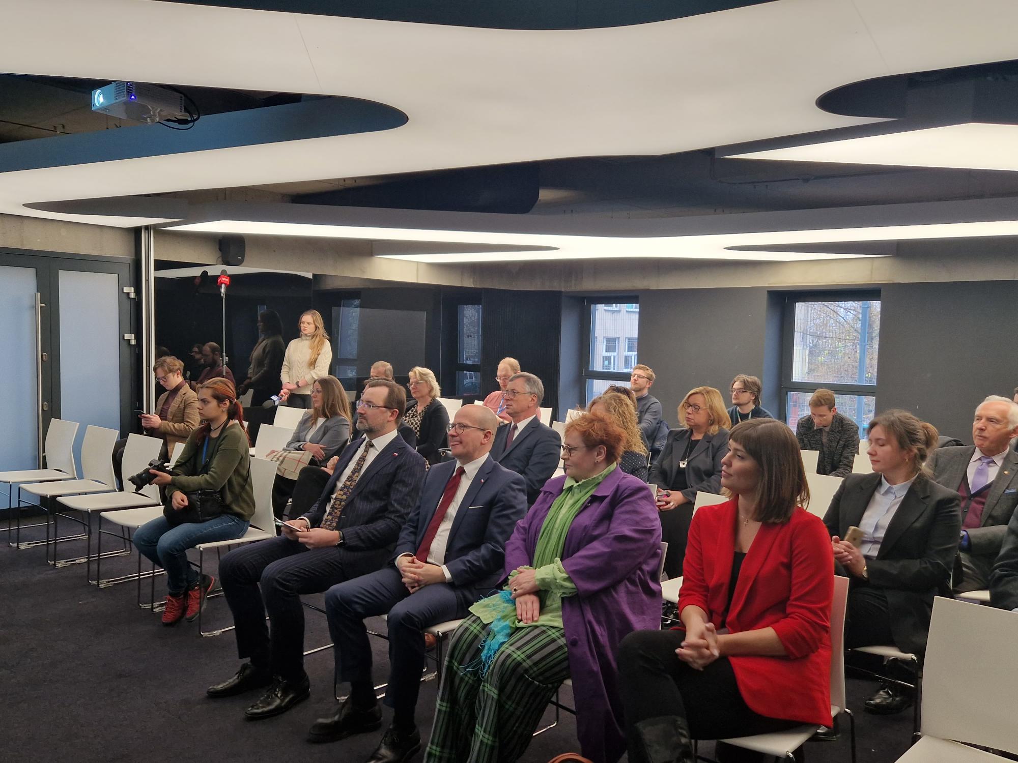 Na zdjęciu znajduje się grupa ludzi, która siedzi na krzesłach w sali.