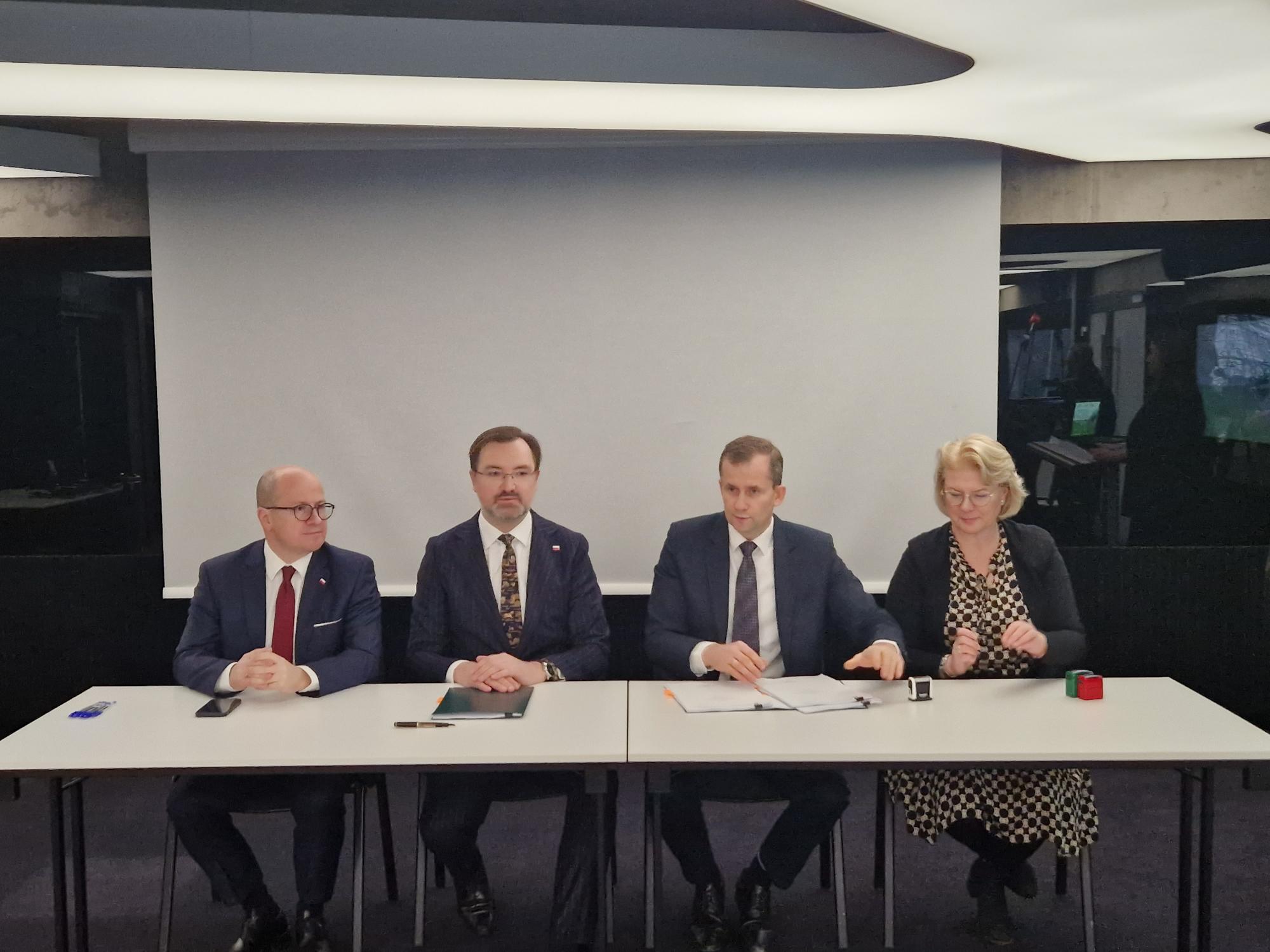 Na zdjęciu 4 osoby siedzą za stołem na tle ekranu.