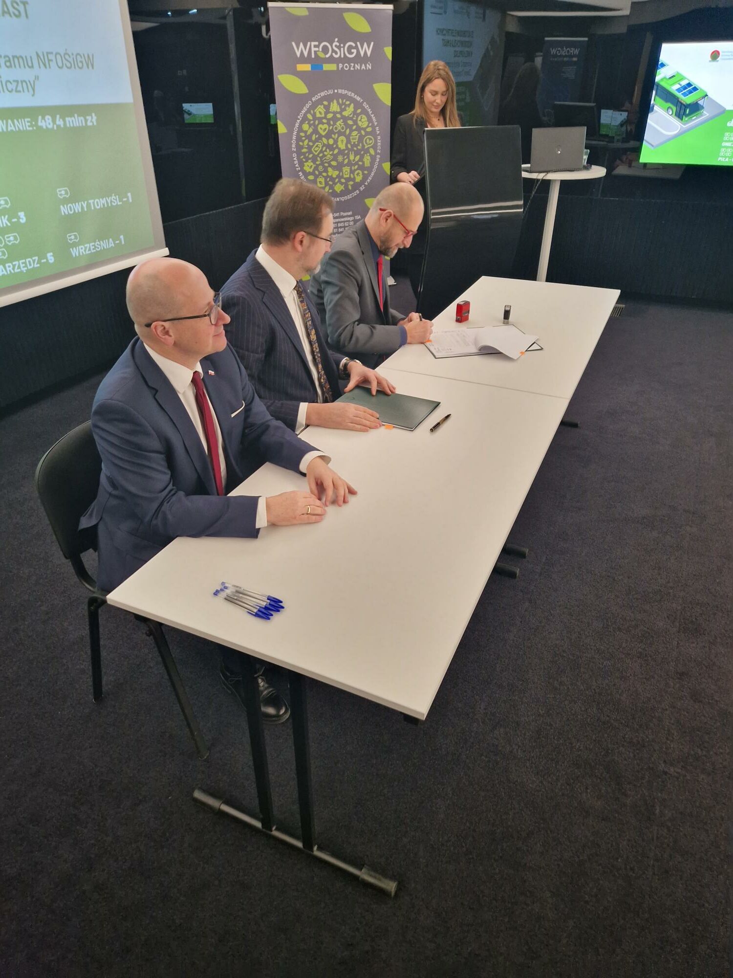 Na zdjęciu 3 osoby siedzą za stołem, a jedna osoba stoi przy mównicy.