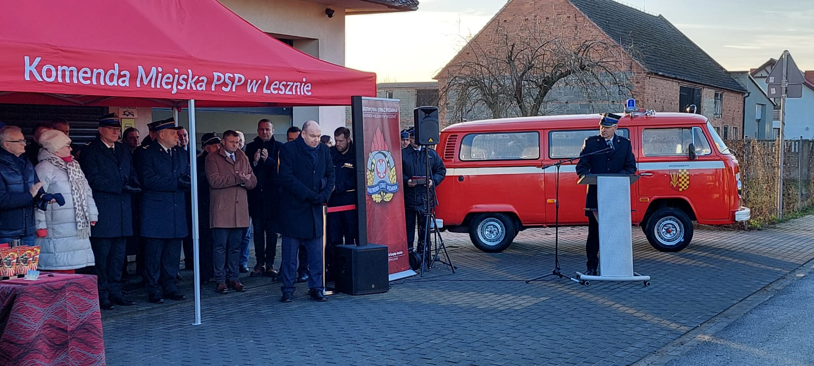 1 osoba stoi przy mównicy na tle samochodu strażackiego