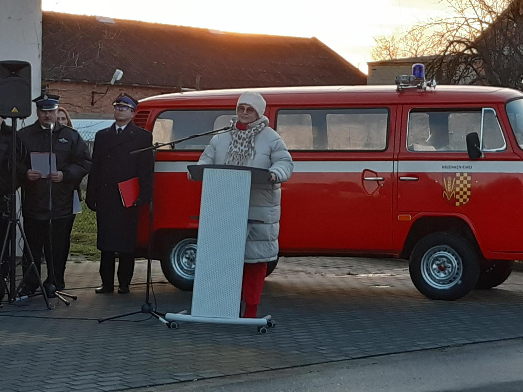 1 osoba stoi przy mównicy na tle wozu strażackiego