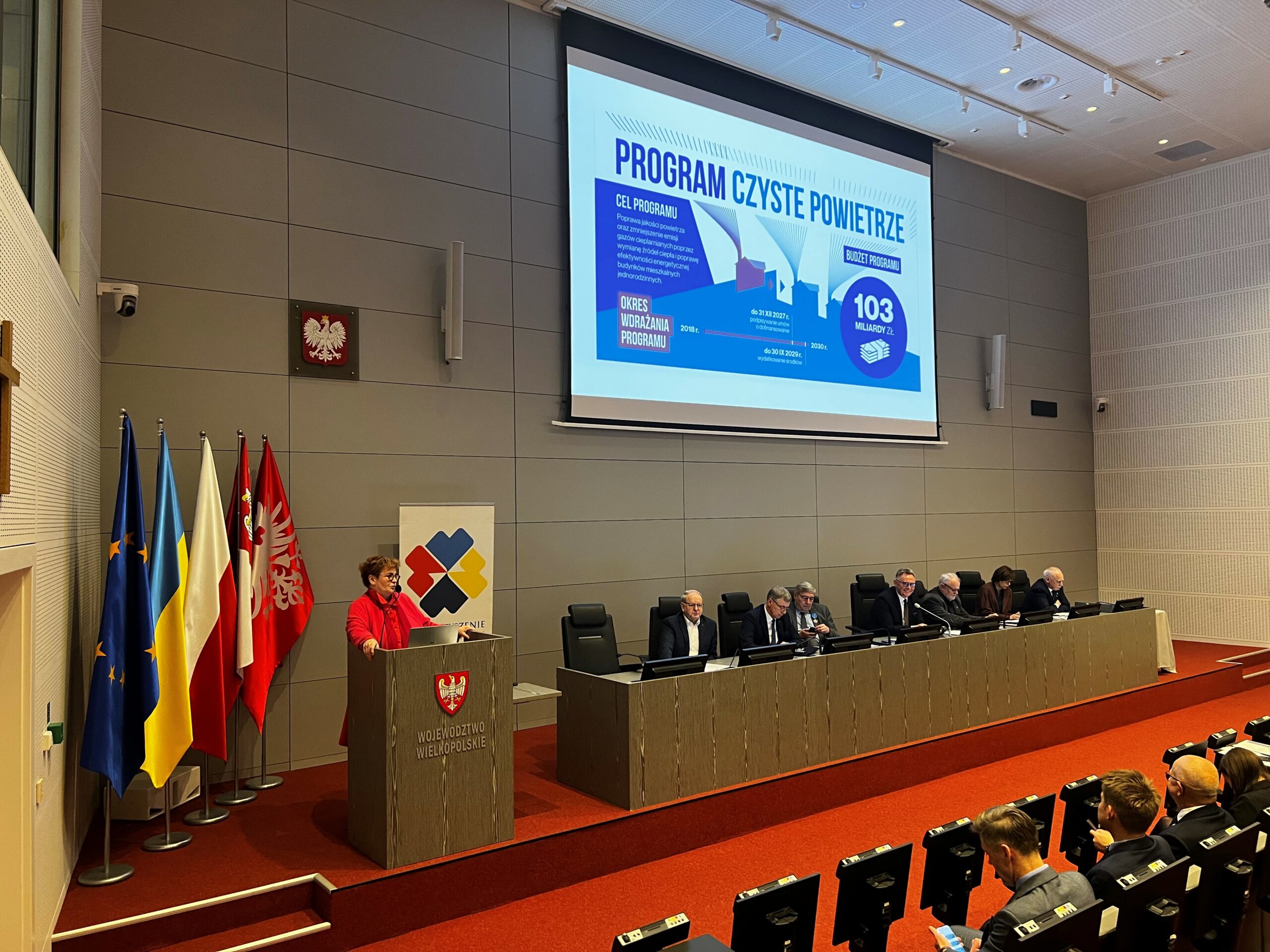 Osoby siedzą przy stole na scenie, a jedna osoba stoi przy mównicy i omawia prezentację.