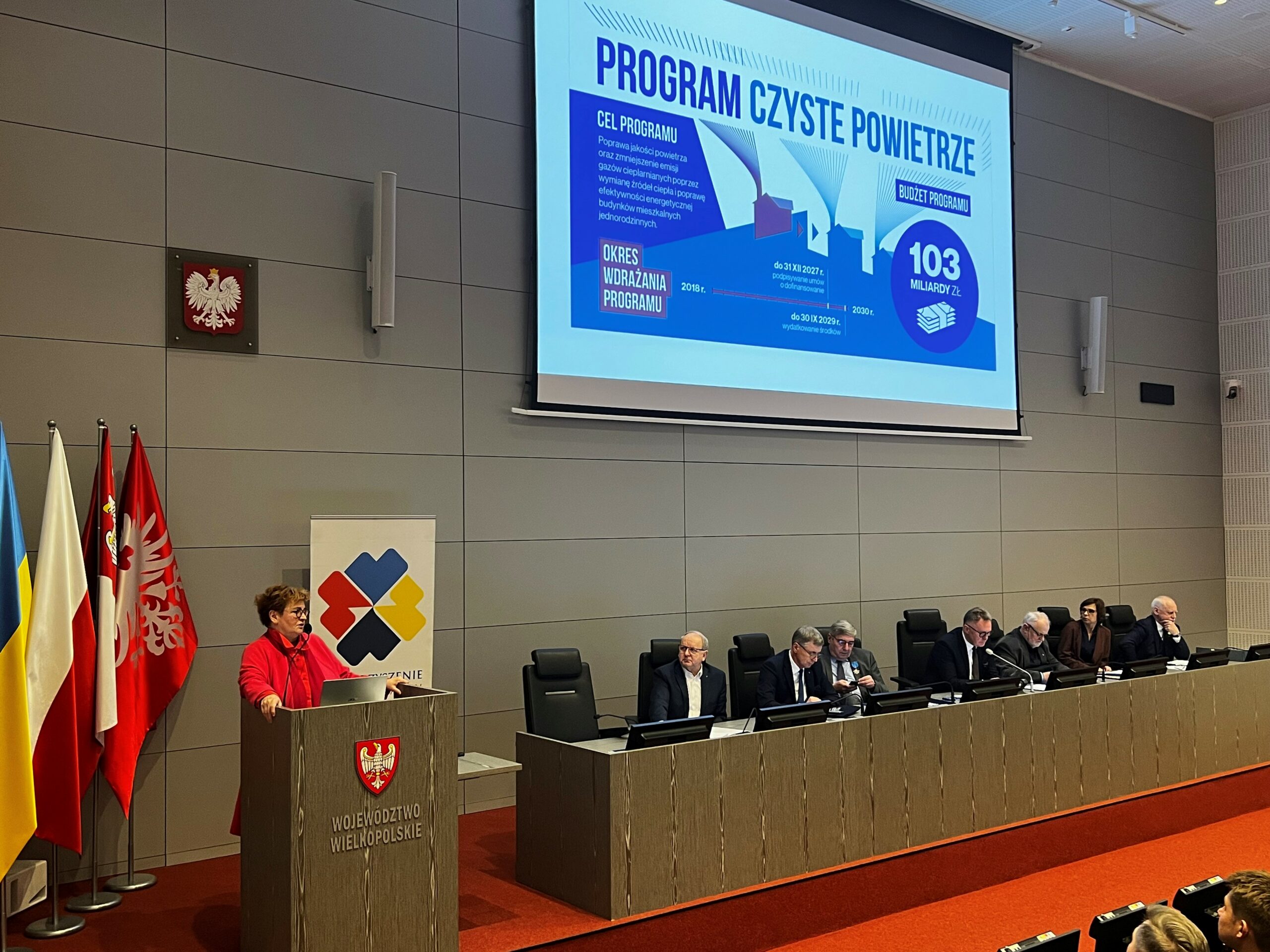 Osoby siedzą przy stole na scenie, a jedna osoba stoi przy mównicy i omawia prezentację.