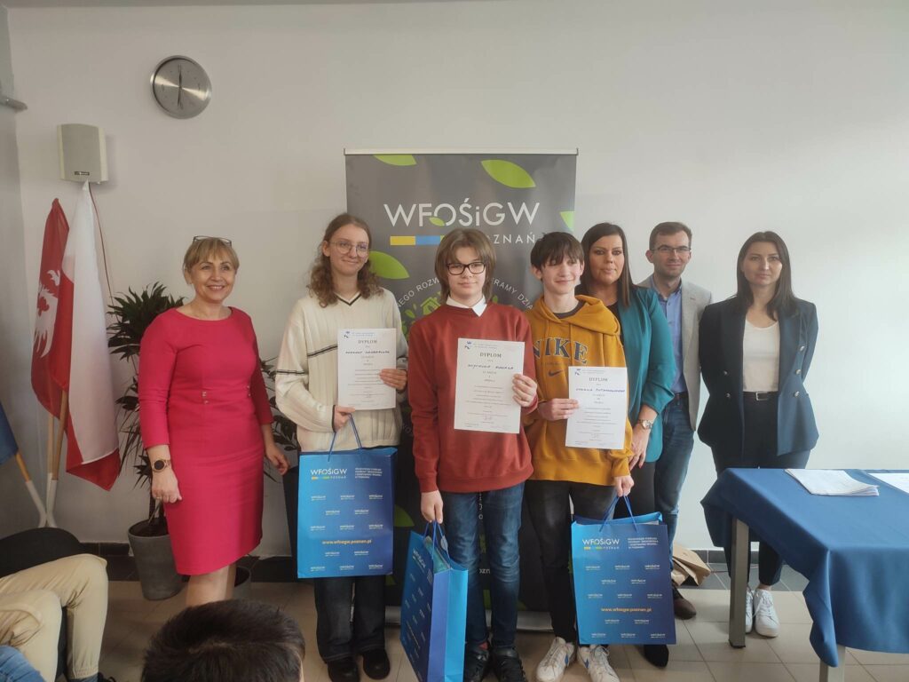 7 osób stoi na tle ściany, jedna wręcza dyplom.