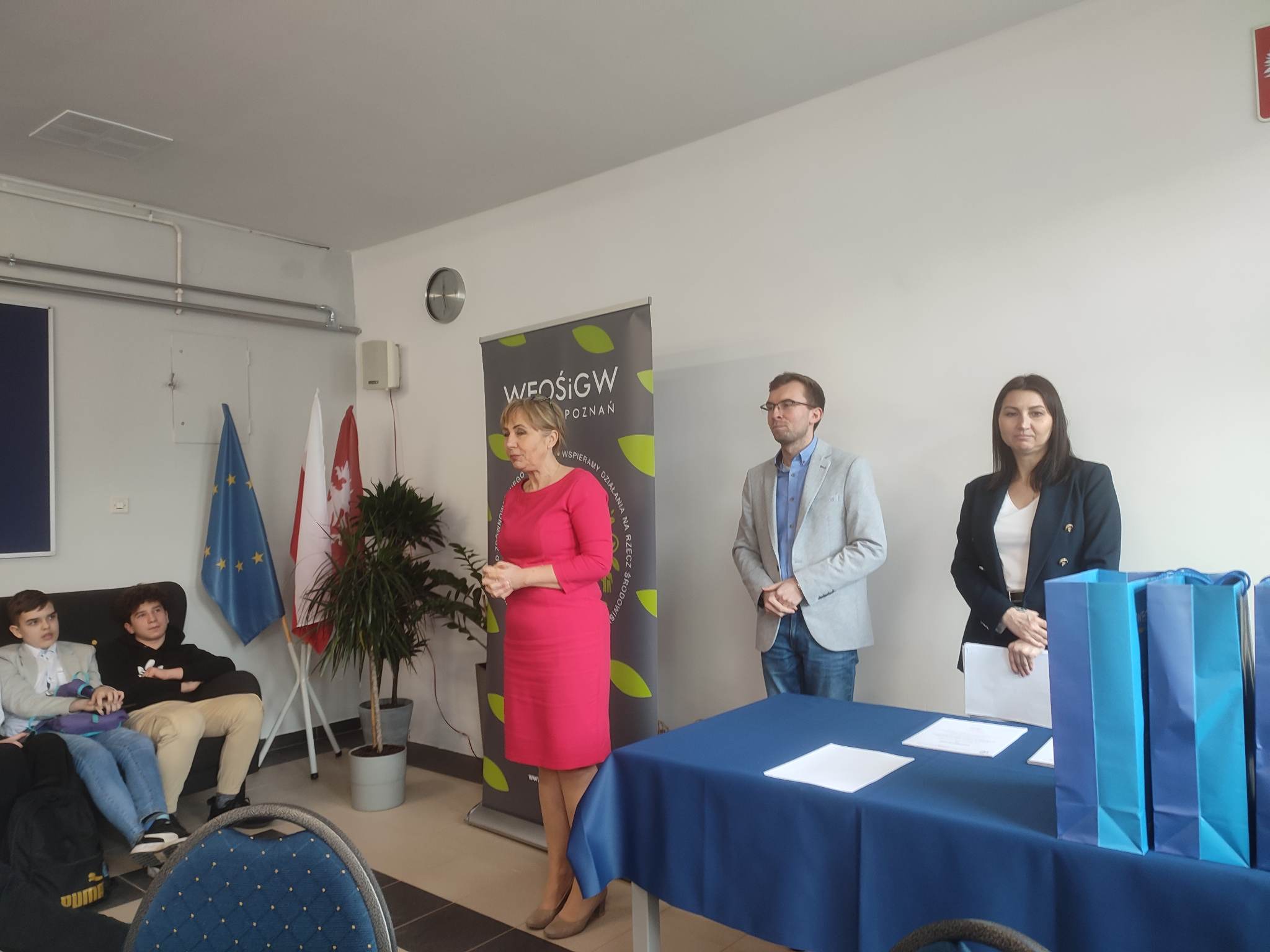 3 osoby stoją w sali na tle ściany.