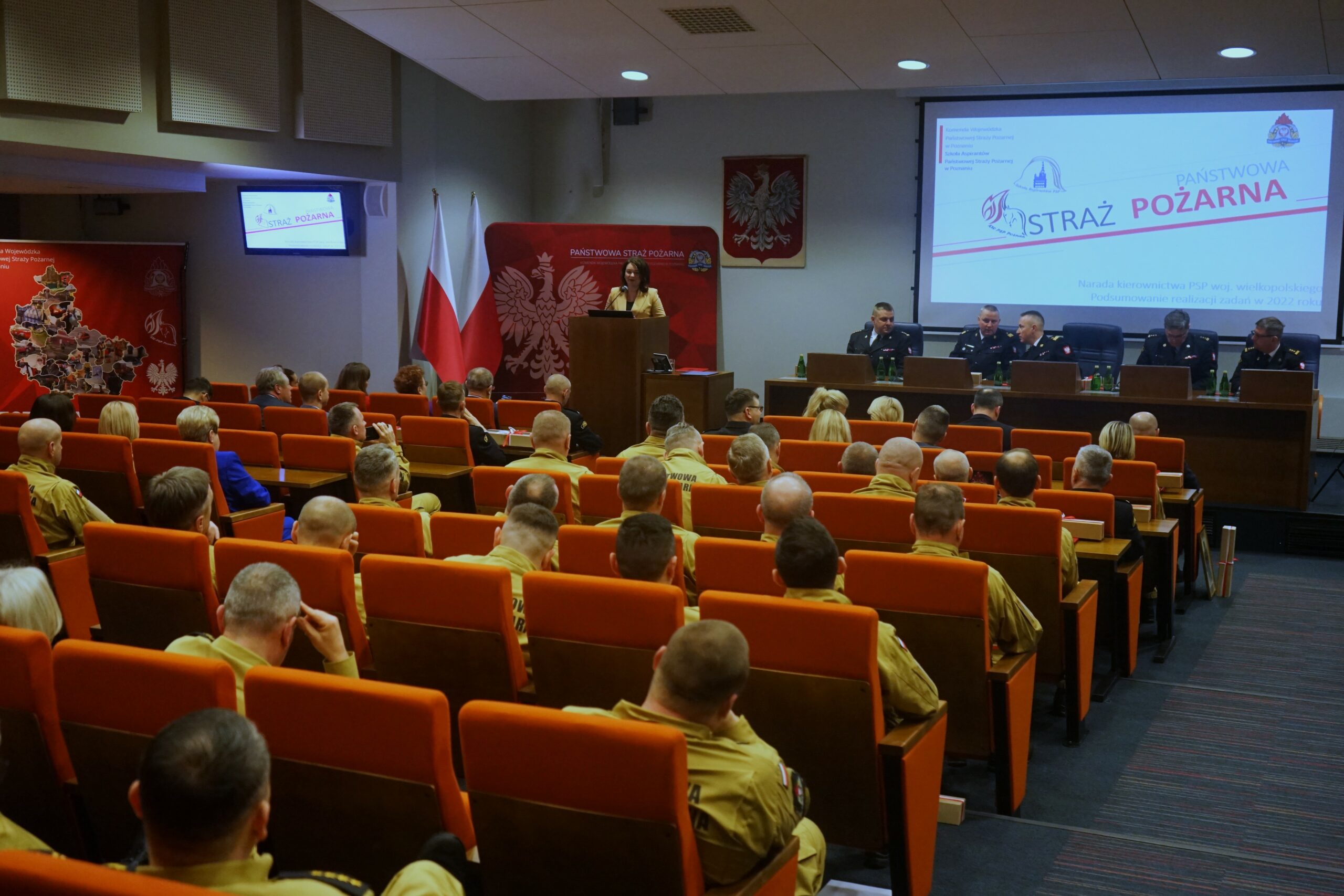 Sala wypełniona ludźmi, przy mównicy stoi jedna osoba i przemawia.