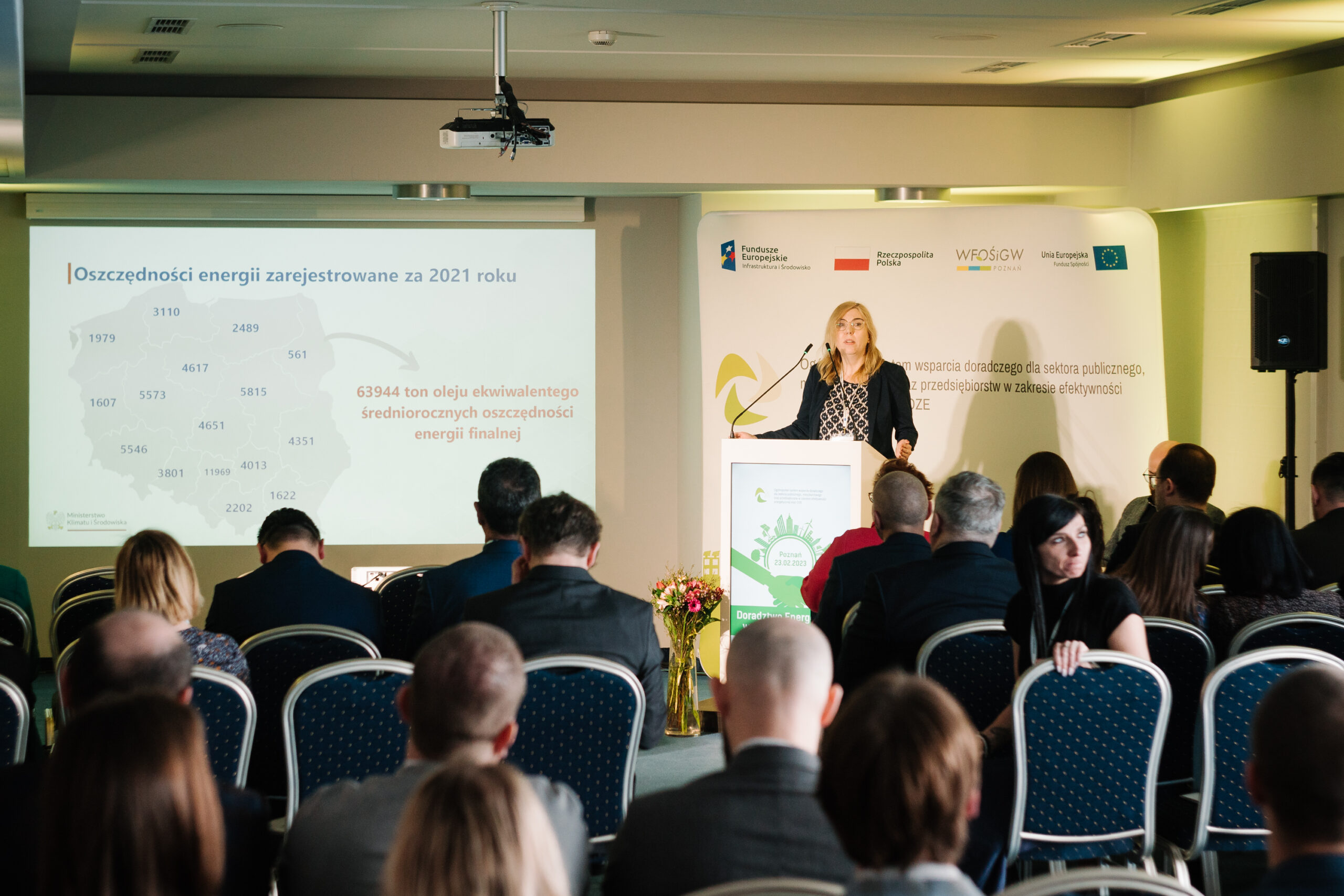 Sala wypełniona ludźmi, a jedna osoba przemawia stojąc przy mównicy.