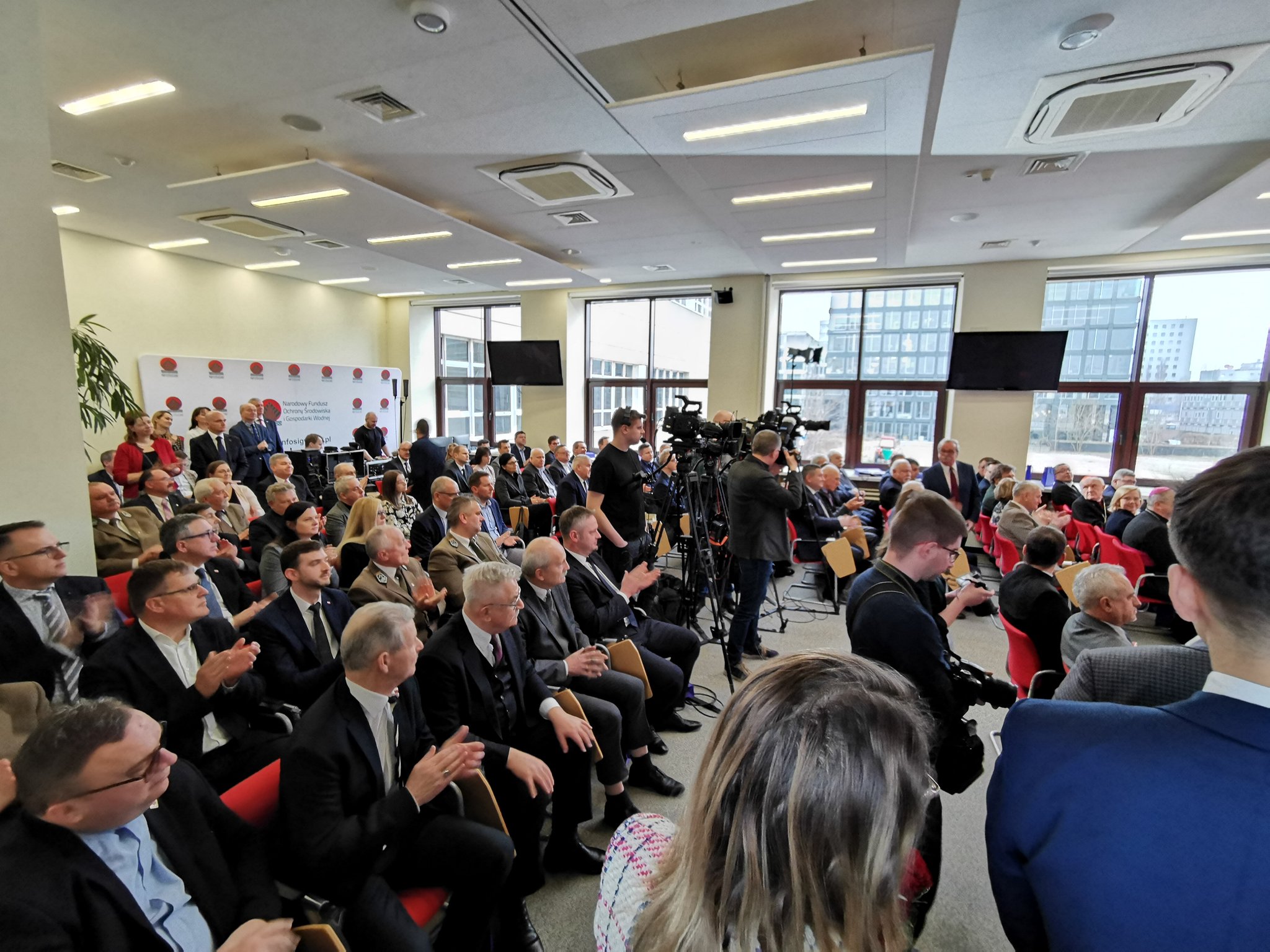 Sala wypełniona ludźmi, którzy siedzą na ustawionych krzesłach.