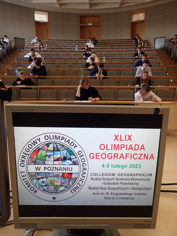 Sala wykładowa a na niej monitor z nazwą wydarzenia "Olimpiada gegraficzna" i siedzącymi w tle uczniami.