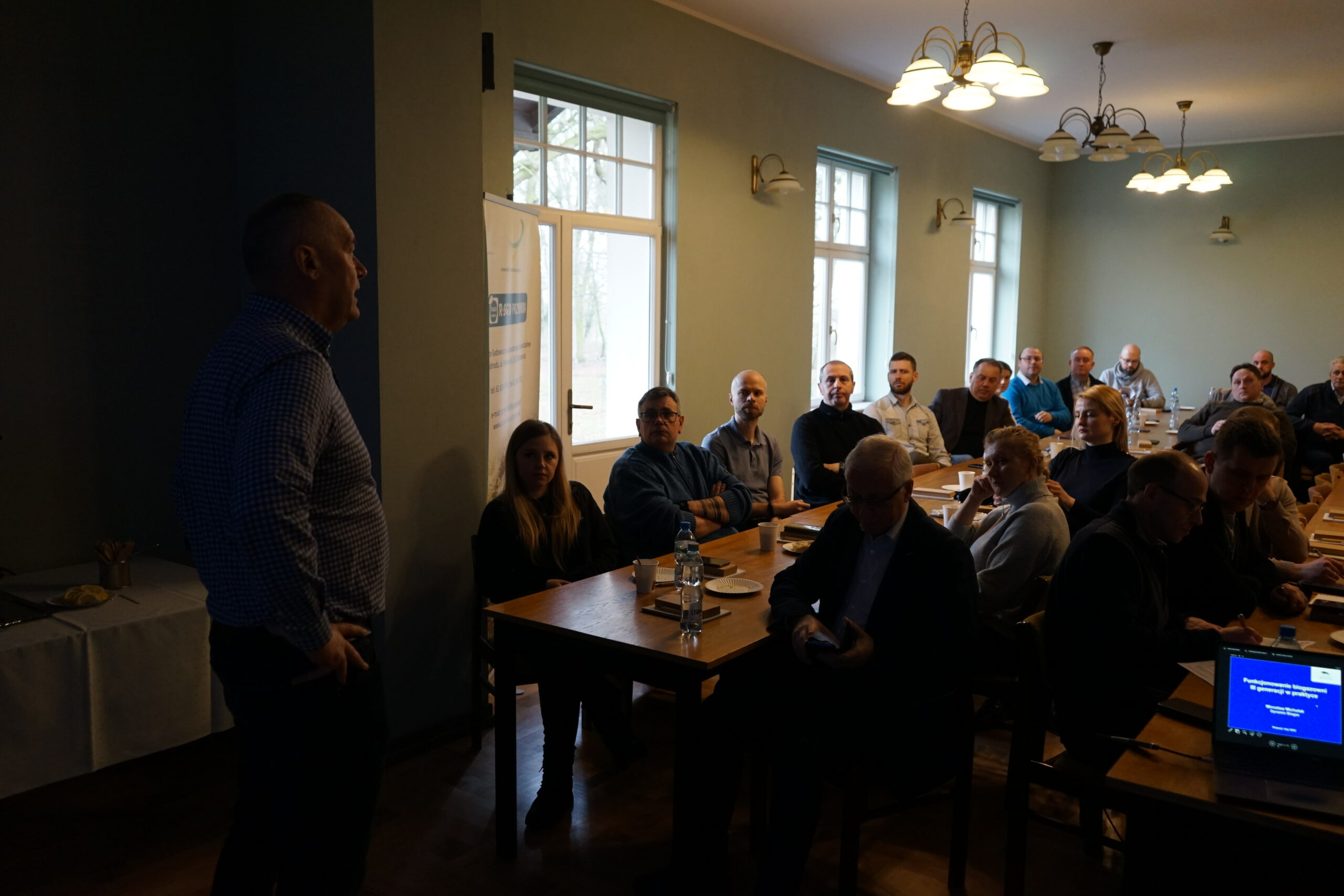 Duża grupa ludzi siedzi na sali i słucha prelekcji patrząc na prezentację.