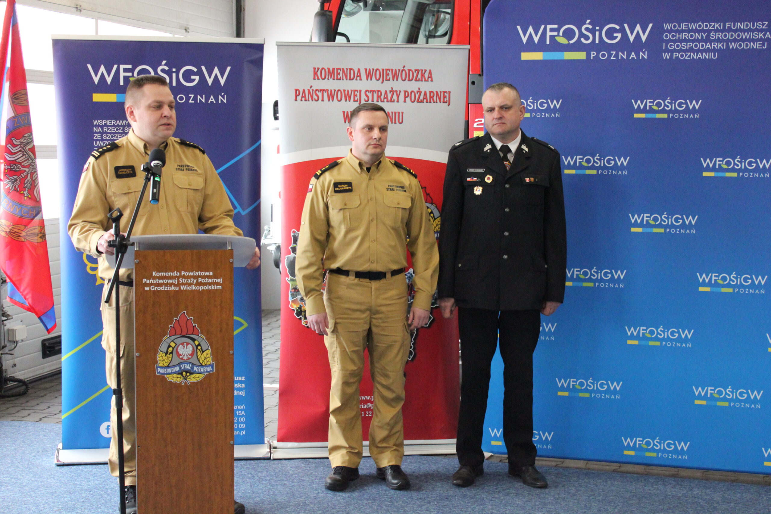 1 osoba stoi przy mównicy, a obok niej stoją jeszcze 2 osoby.