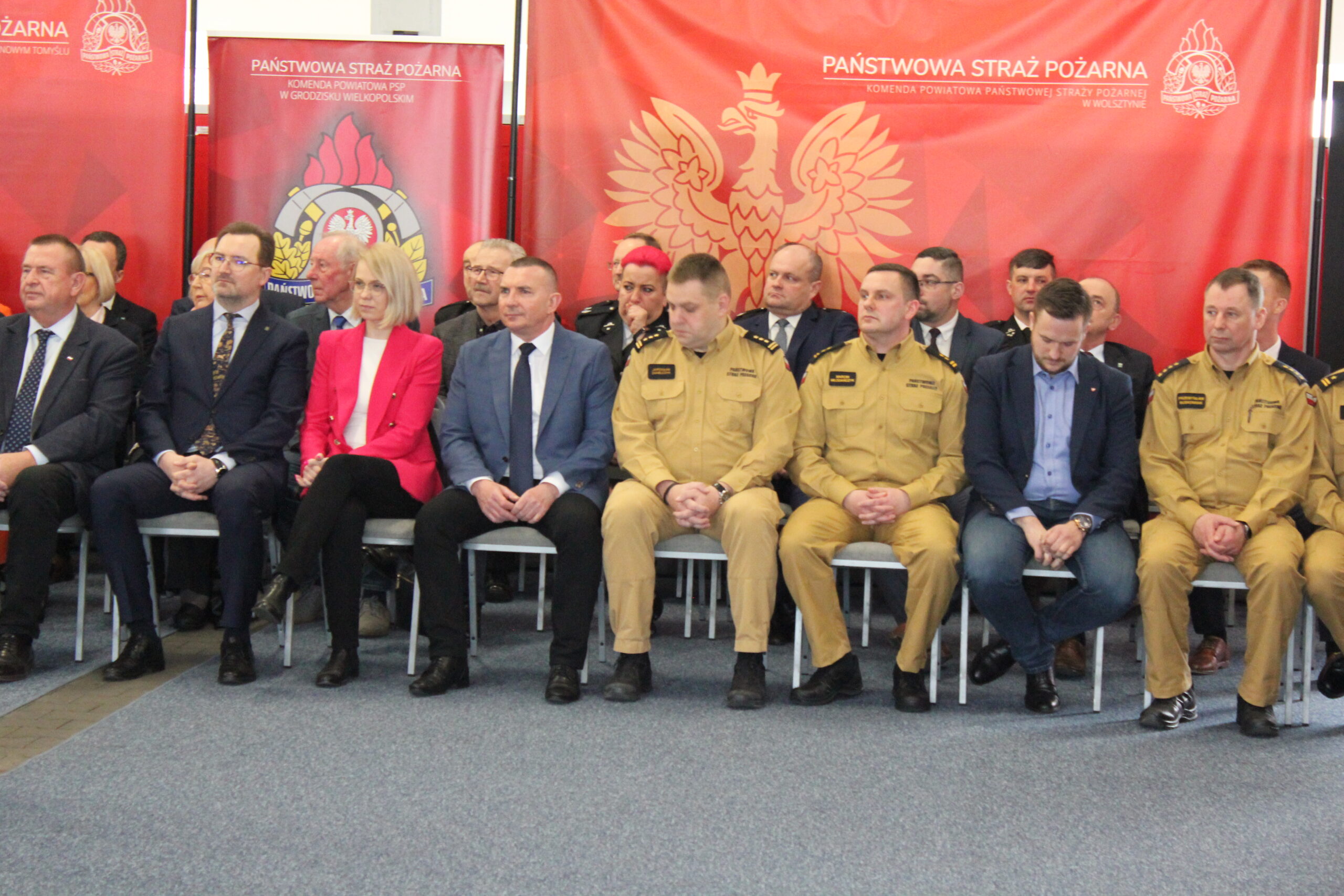 Dużo osób siedzi na krzesłach na tle bannerów reklamowych.