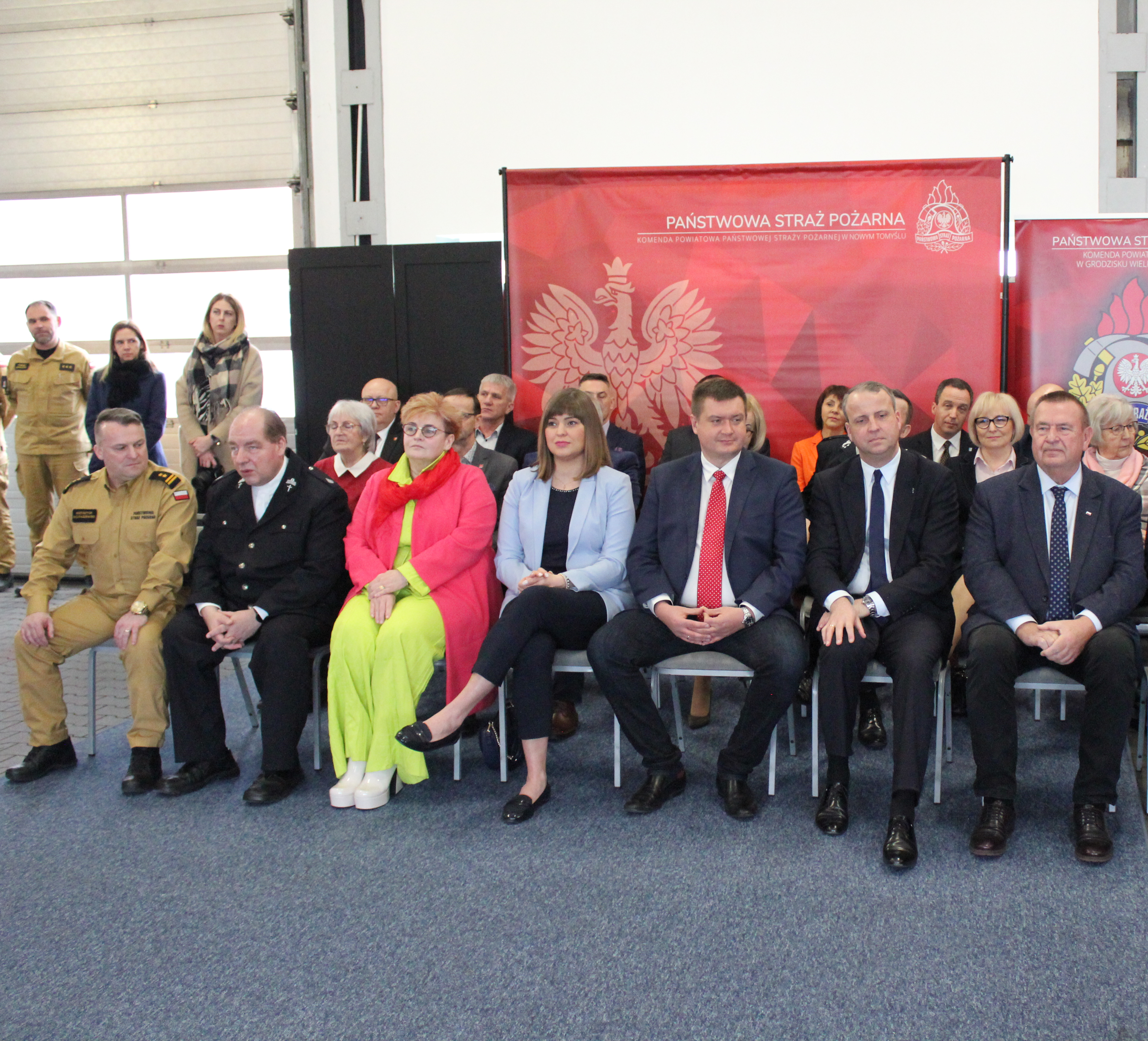 Duża grupa osób siedzi na krzesłach na tle bannerow reklamowych.