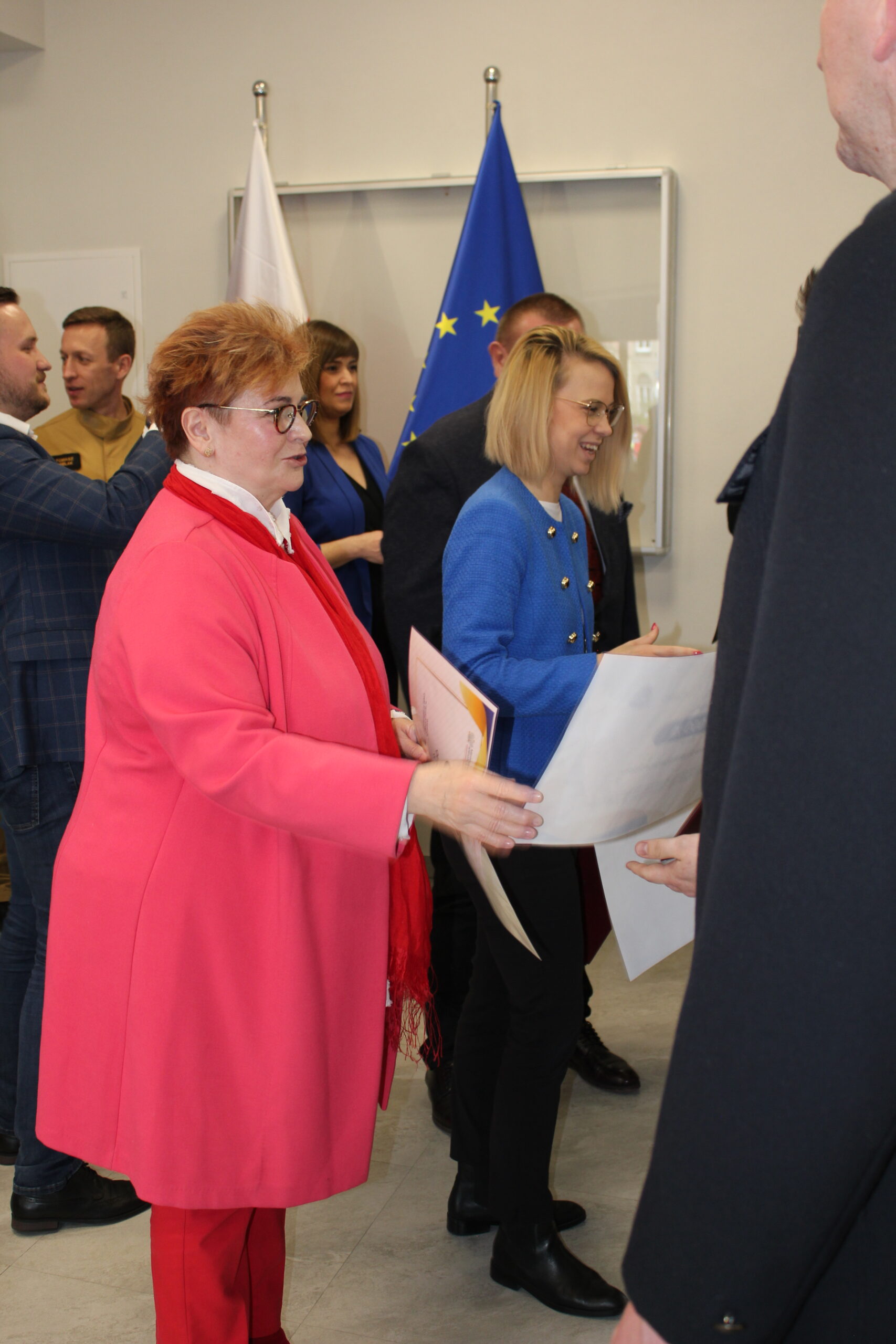 Osoby w rzędzie stoją na sali i są im wręczane promesy.