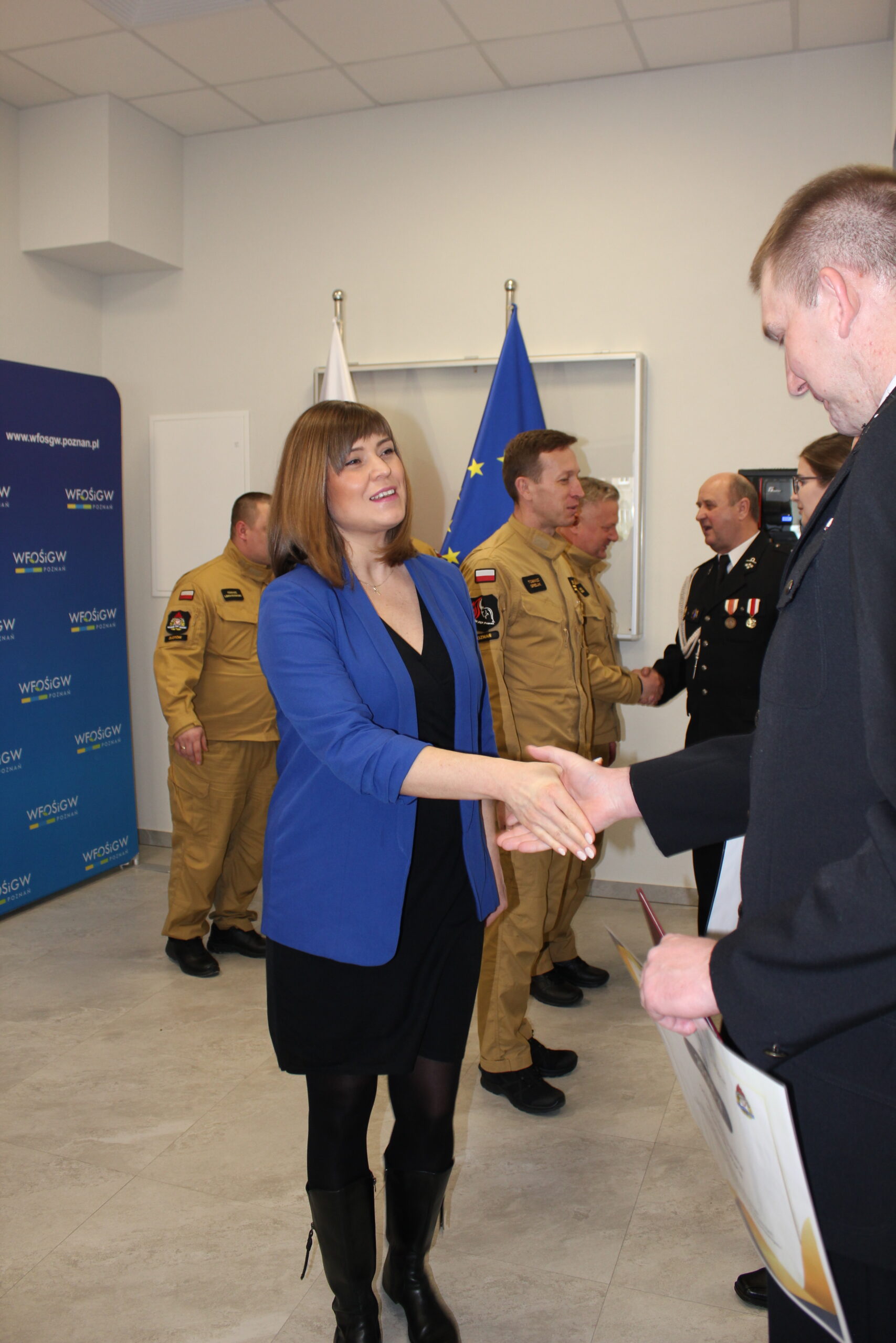 Osoby w rzędzie stoją na sali i są im wręczane promesy.