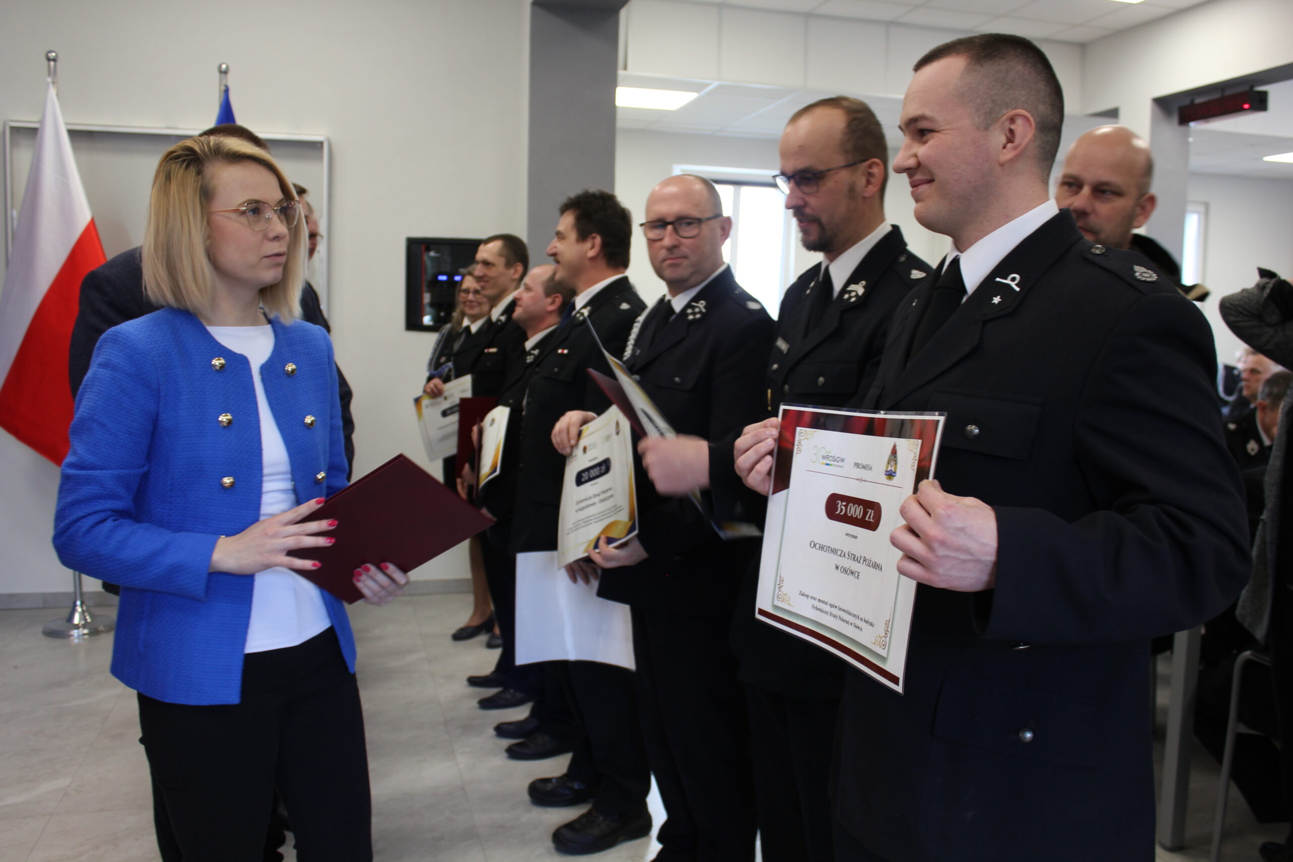 Osoby w rzędzie stoją na sali i są im wręczane promesy.