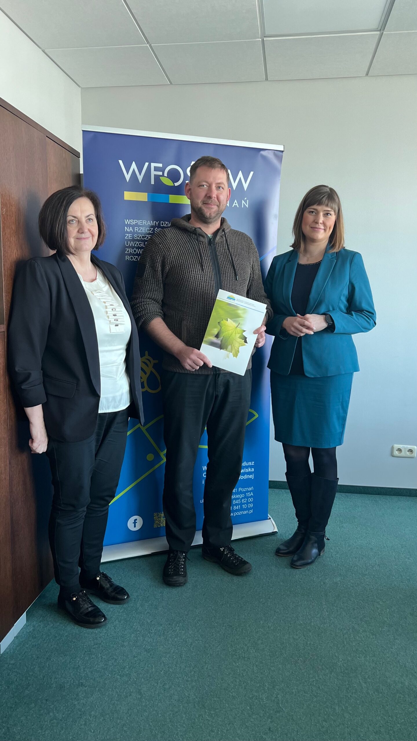 3 osoby stoją na tle ściany i bannera reklamowego.